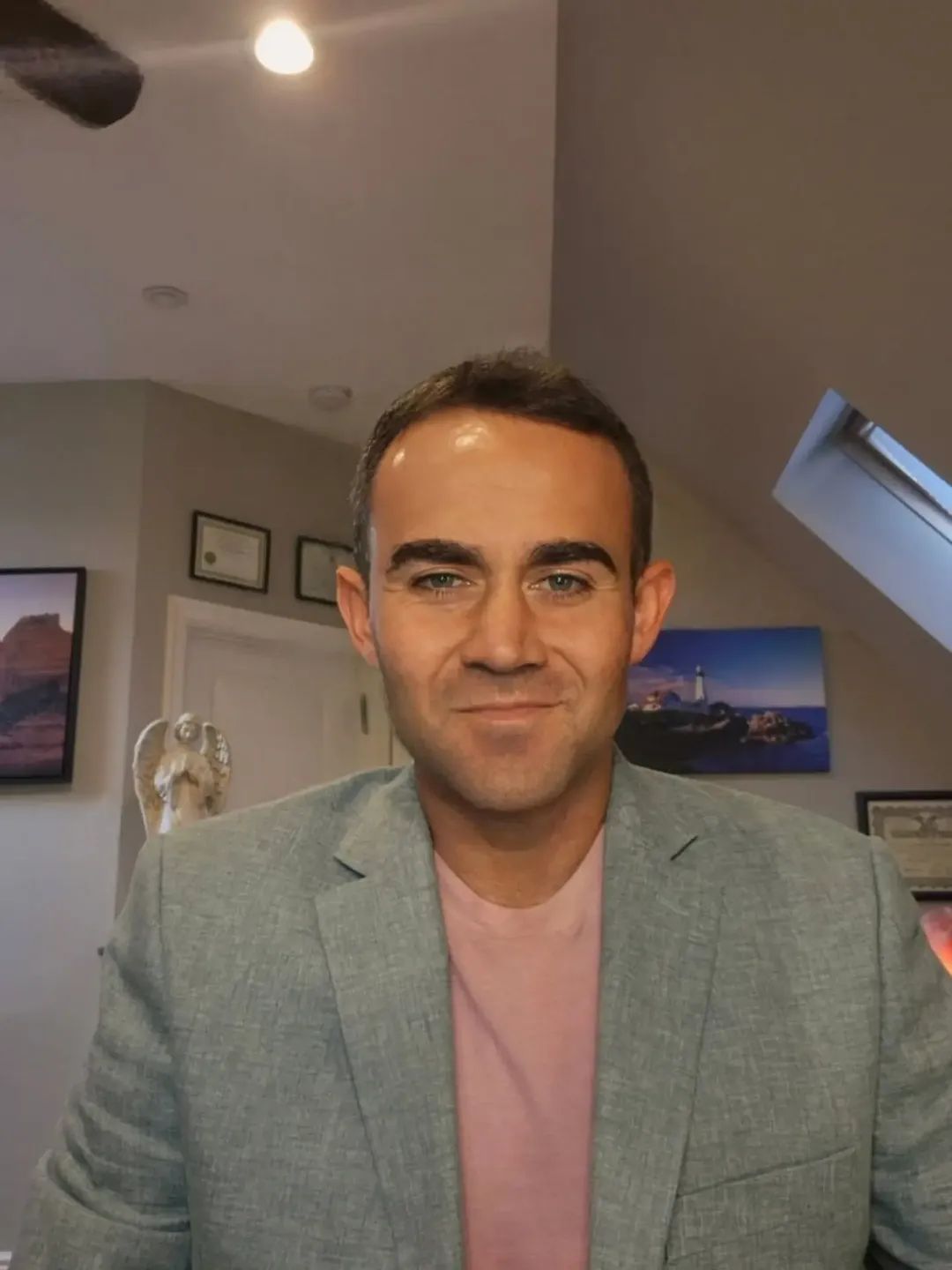 Man with short dark hair and a slight smile, wearing a light gray blazer and pink shirt, indoors with artwork on walls.
