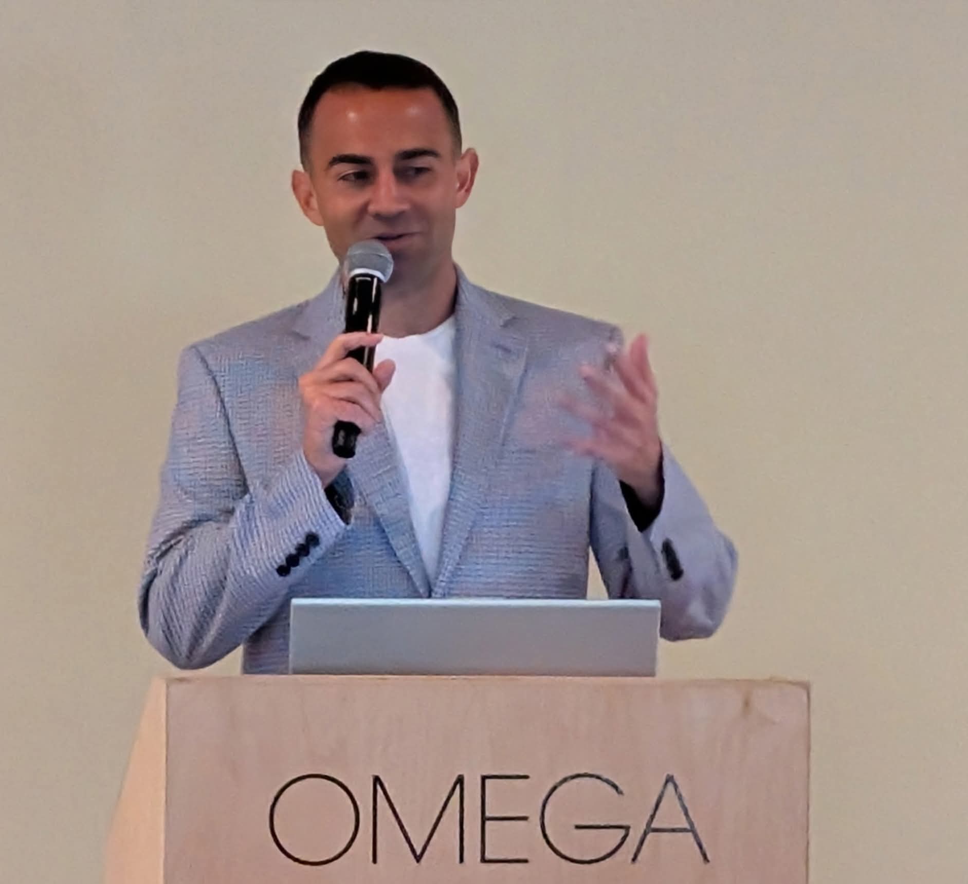 Man speaking into a microphone at a podium with a laptop, in front of a plain background.