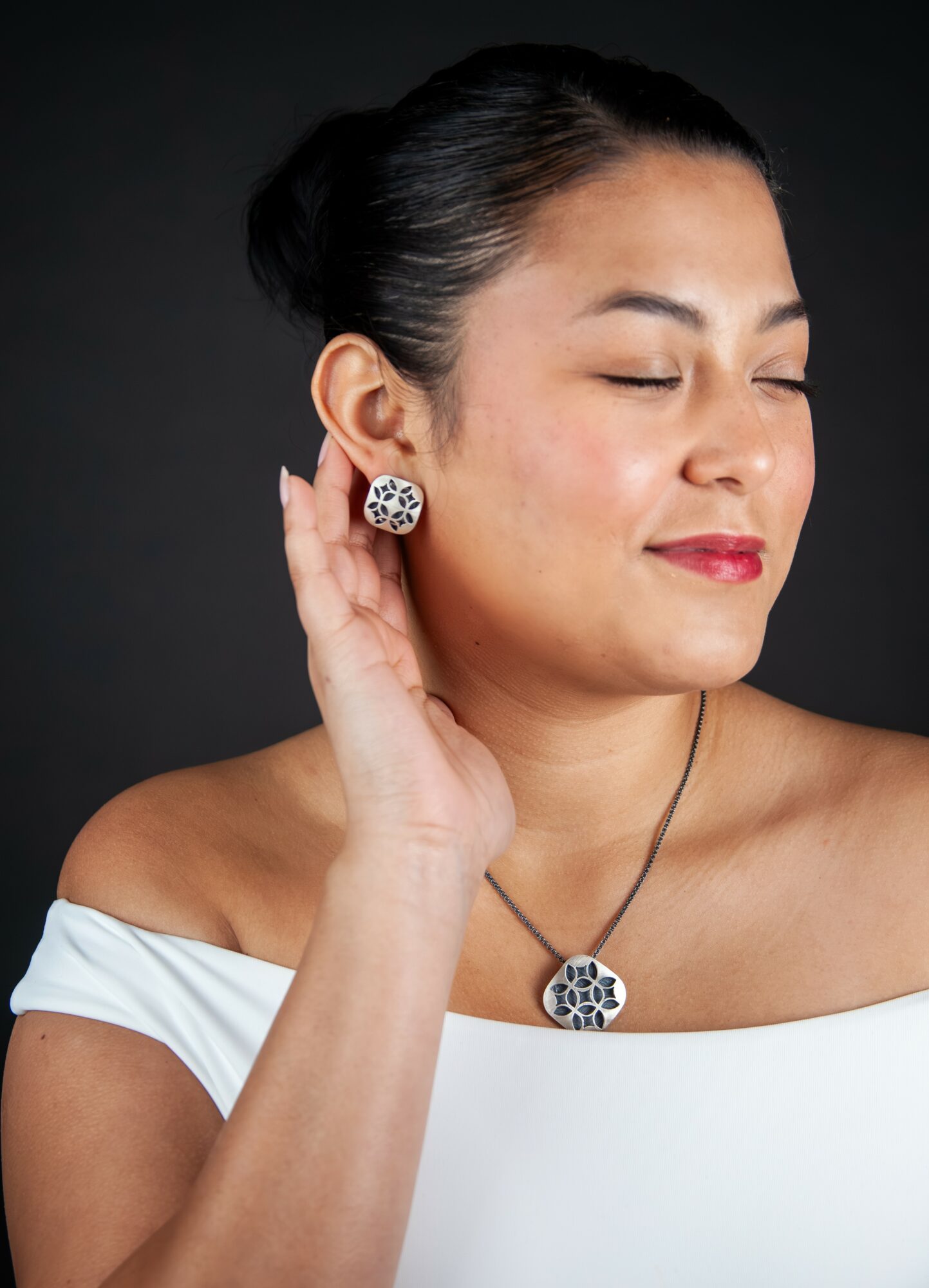 Woman with closed eyes, wearing earrings and a necklace, touching her ear, against a dark background.