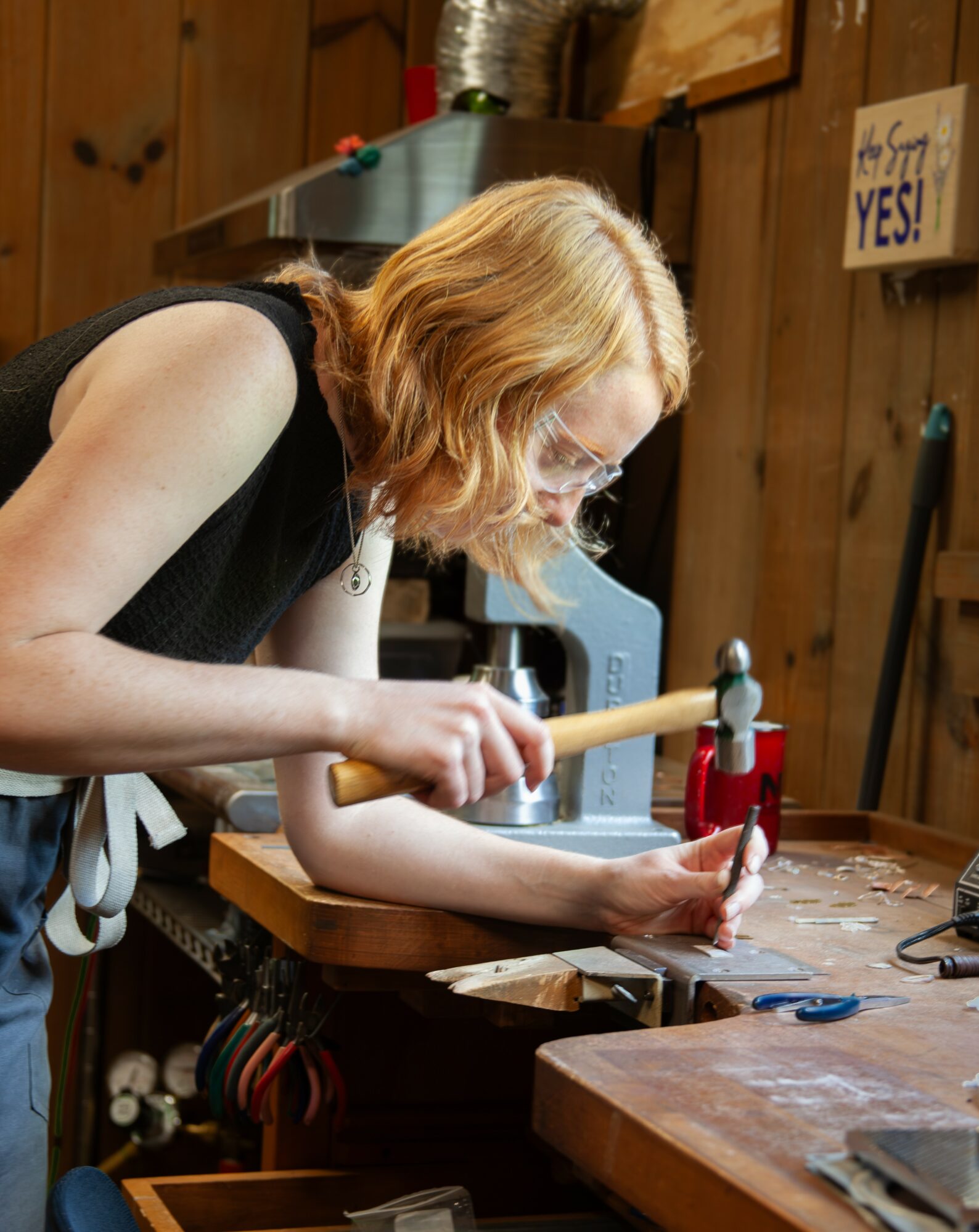 Woman with red hair working with a hammer on a wooden workbench in a workshop.