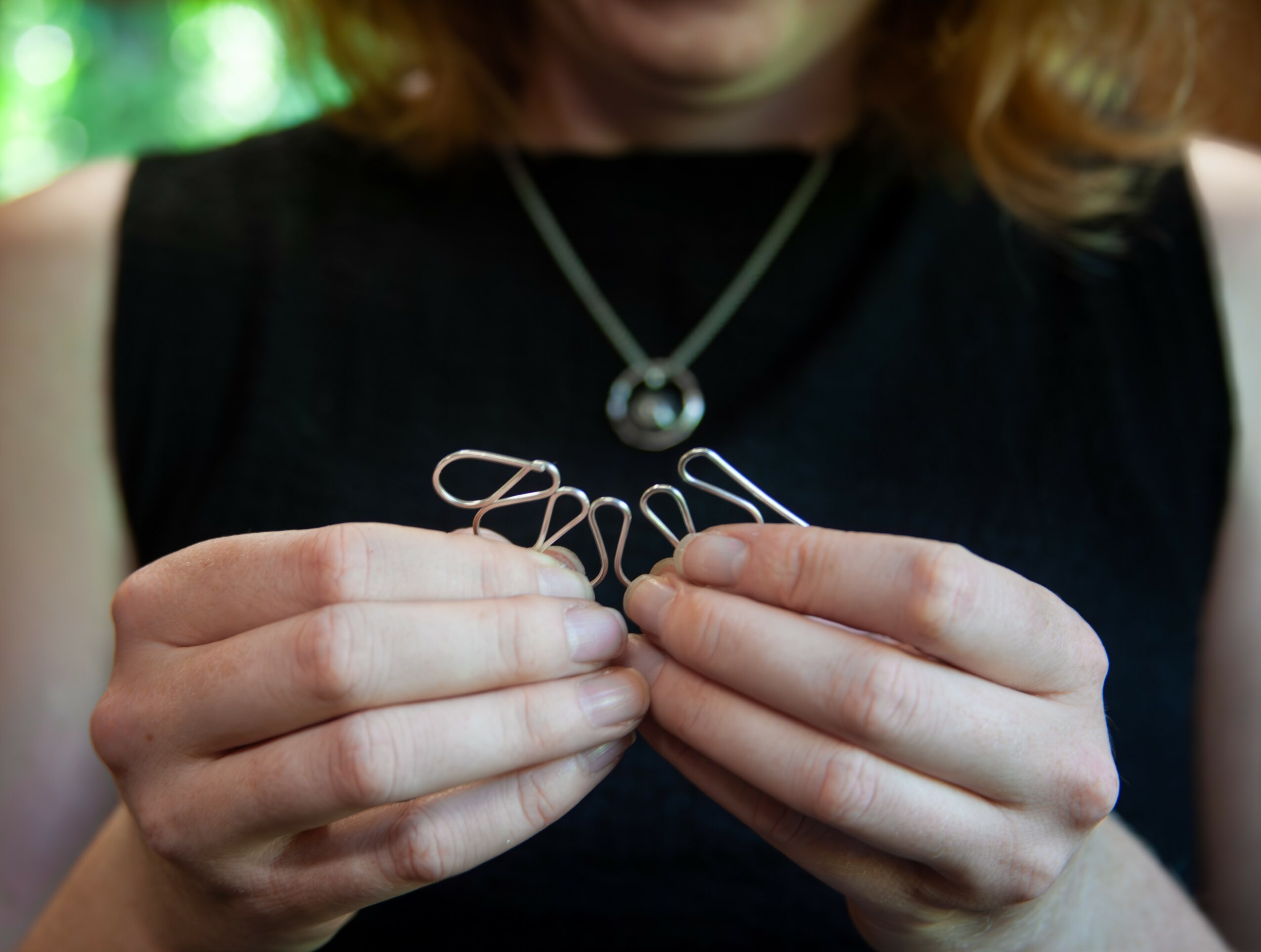 Person holding a delicate wire jewelry piece with both hands, wearing a black sleeveless top and a necklace with a round pendant.