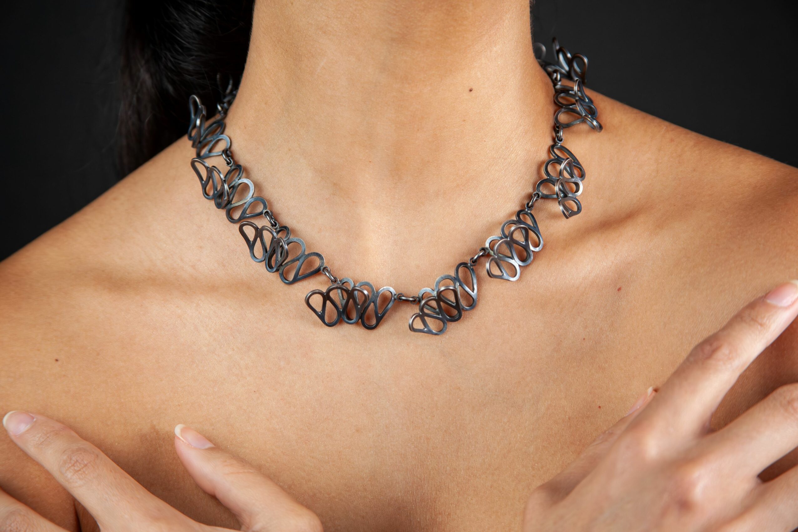 Close-up of a person's neck and shoulders wearing a black, intricate chain necklace, with hand resting on shoulder.