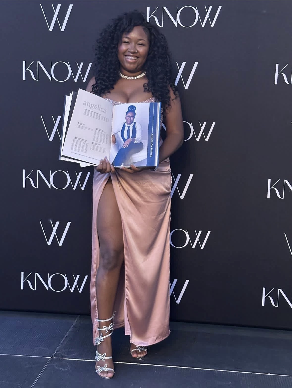 Woman with curly hair holding an open book, standing in front of a black backdrop with white text, smiling.