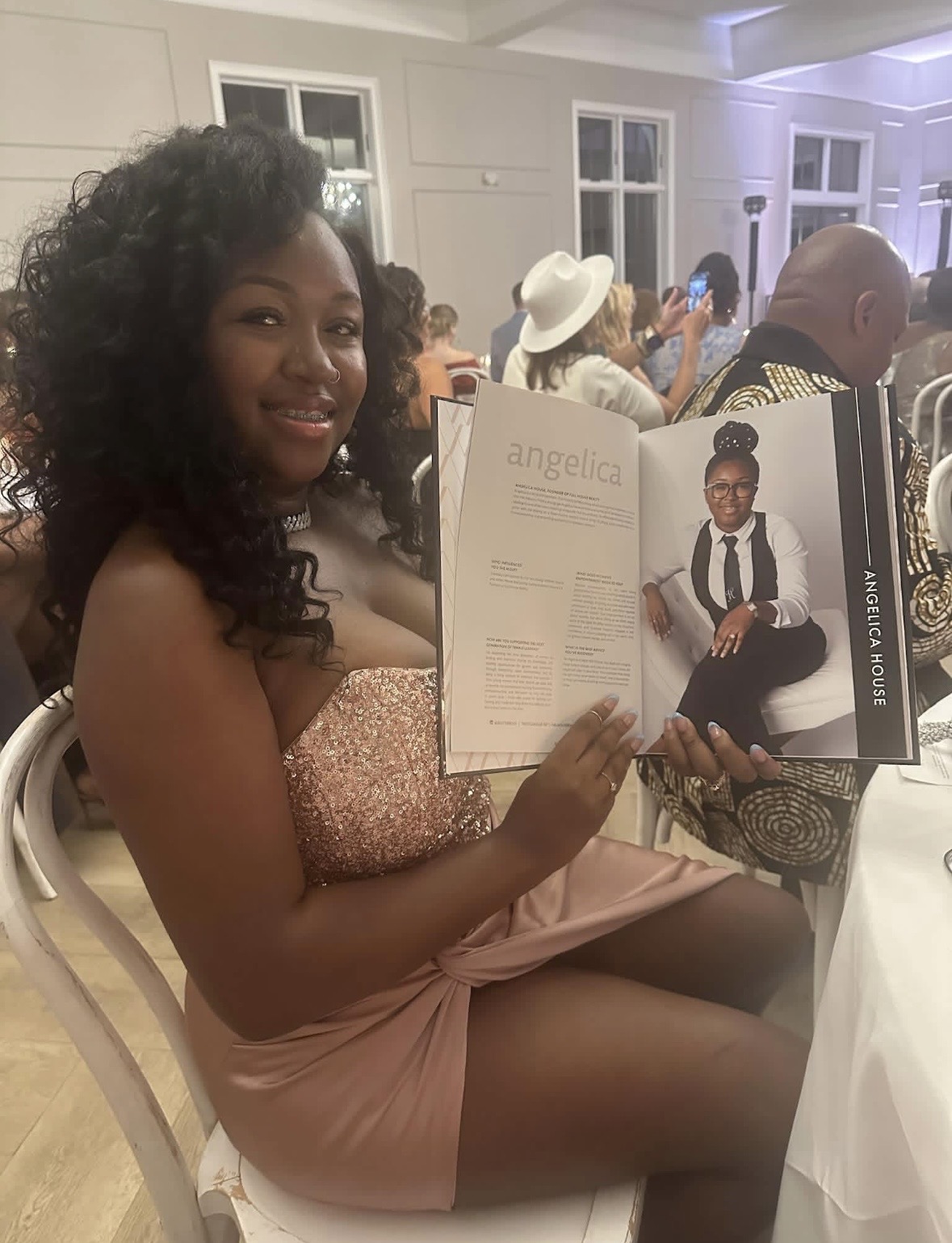 Young woman with curly hair sitting at a table, holding a photo album, smiling at the camera, in a decorated indoor event space.
