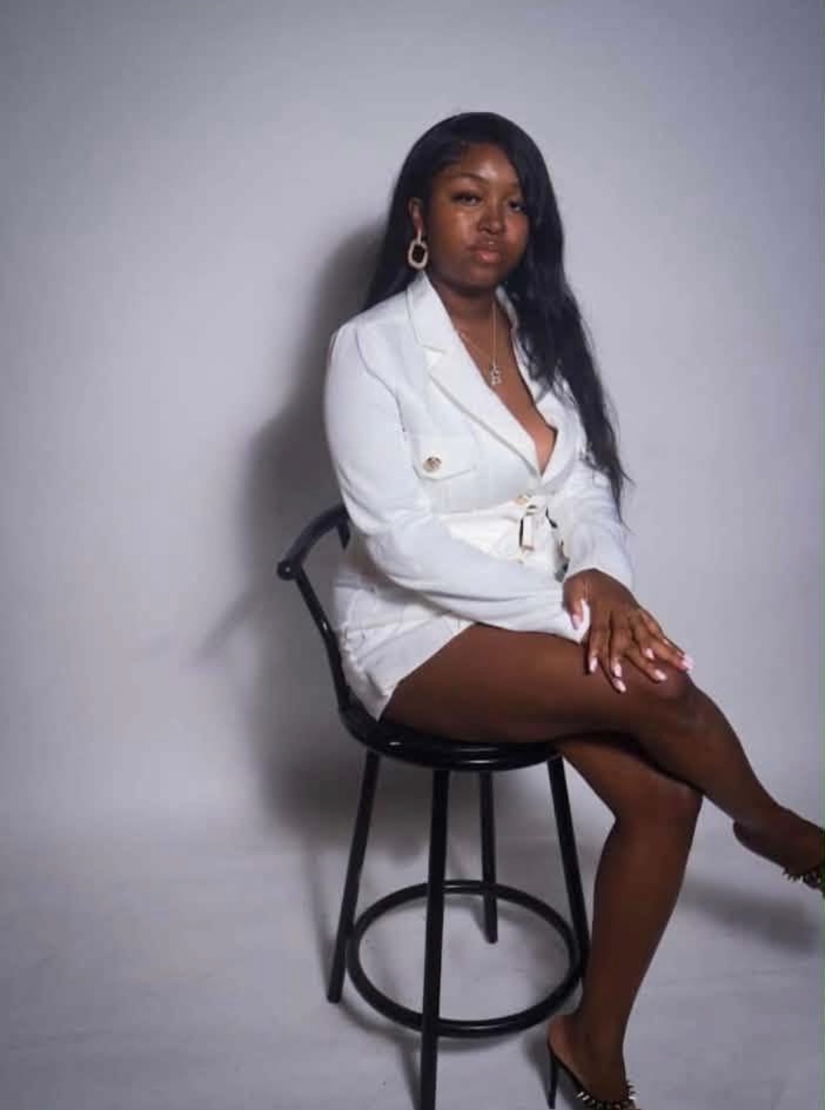 Woman sitting on a black stool, wearing a white blazer and shorts, with long black hair and hoop earrings, against a plain gray background.