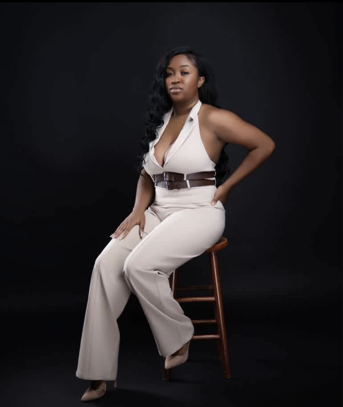 Woman sitting on a wooden chair against a dark background, wearing a white sleeveless top and beige pants, looking at the camera.