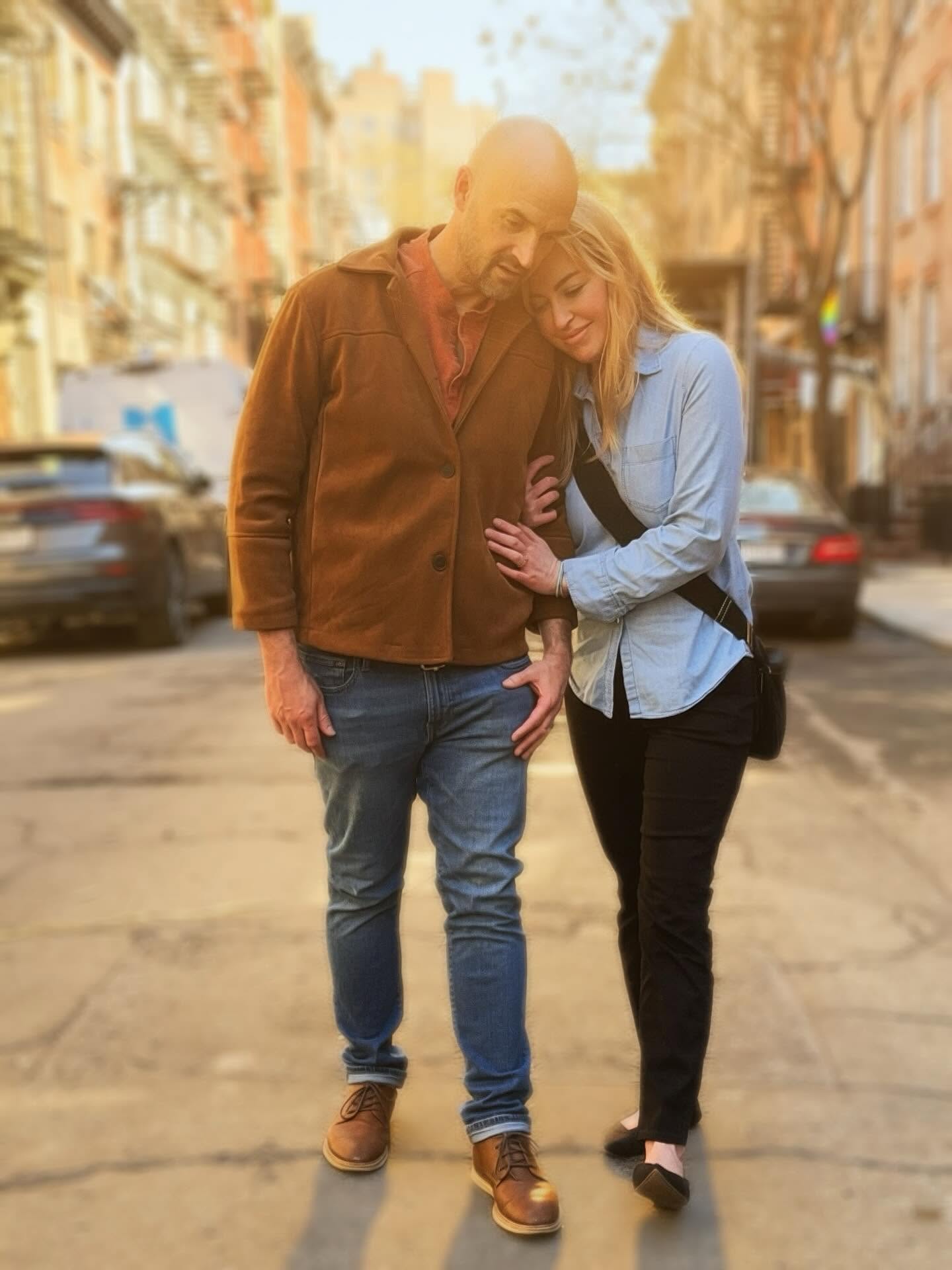 A man and woman walk closely together on a city street, with buildings and parked cars in the background.
