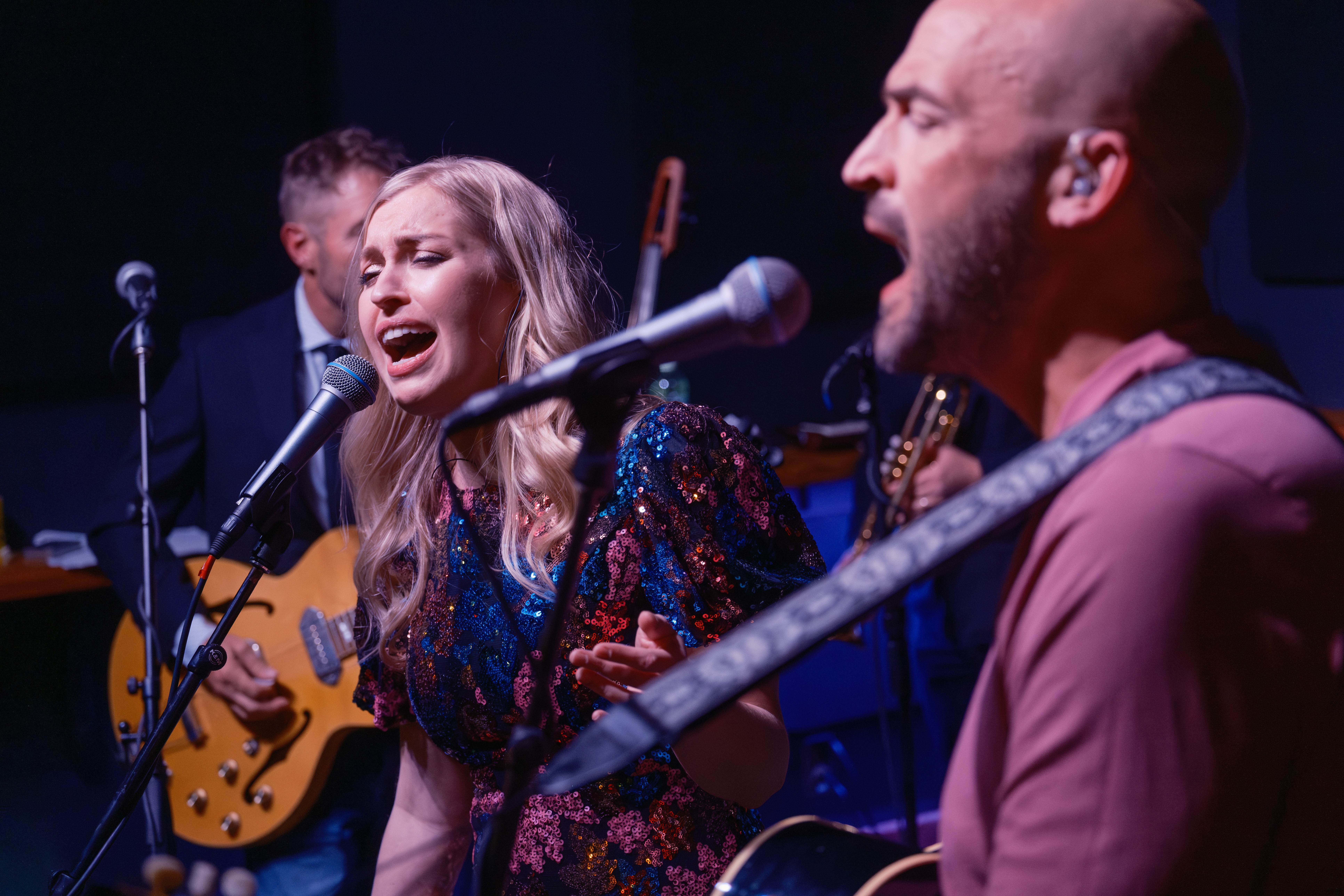 Group of musicians performing on stage, singing and playing instruments, with a woman singing into a microphone.