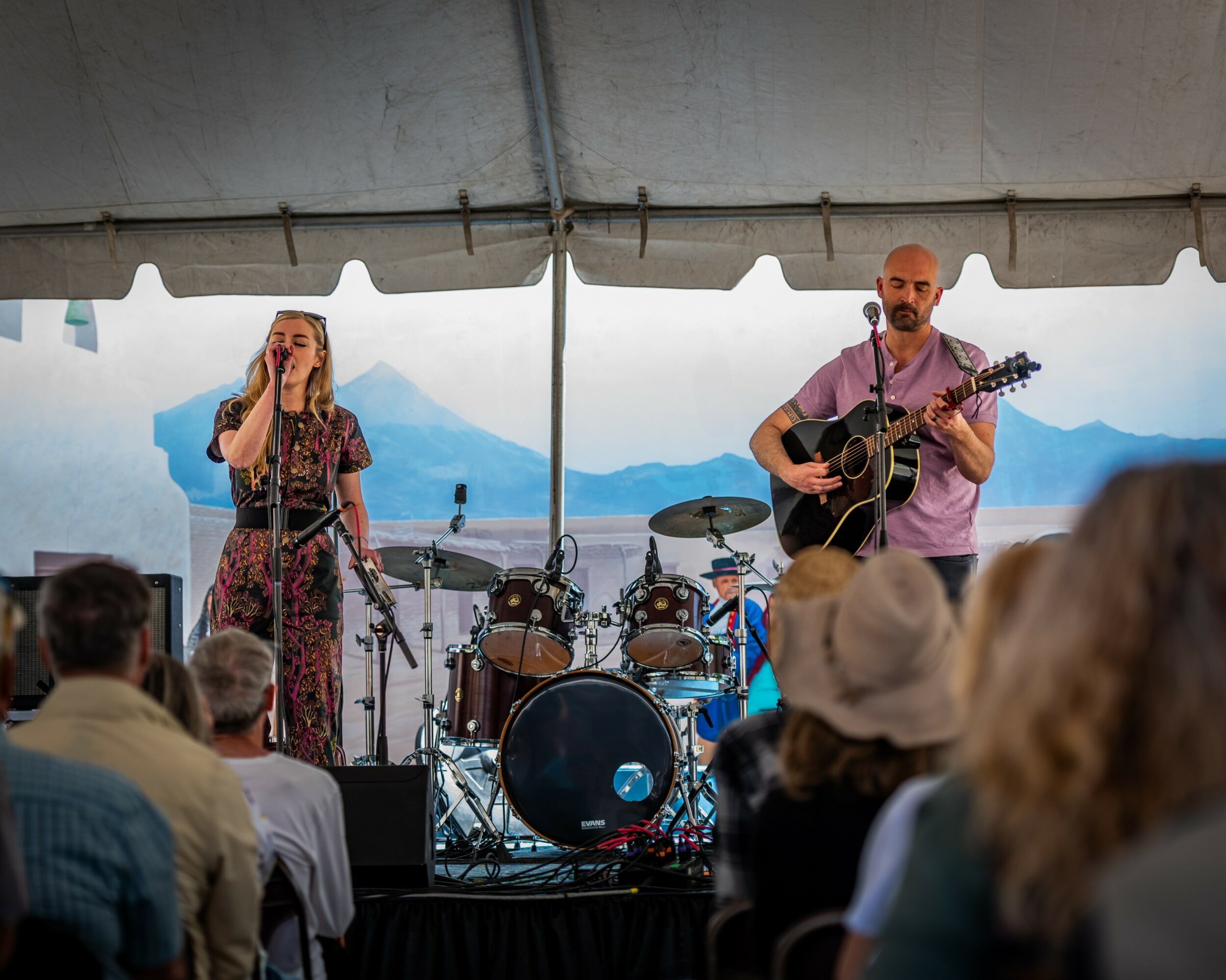 Two musicians perform on stage under a tent, with a drum set and audience in front, mountains in background.