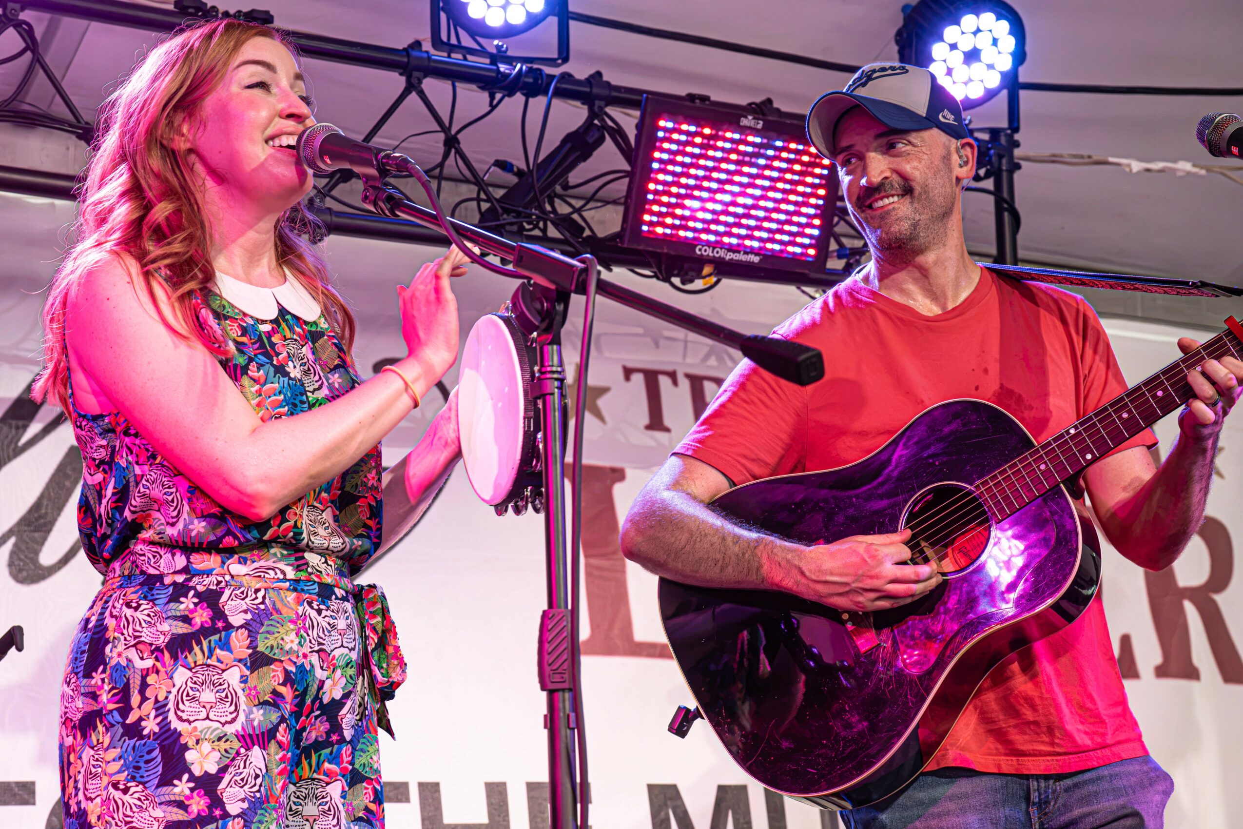 A woman and a man perform on stage, woman singing into a microphone, man playing guitar, stage lights behind them.