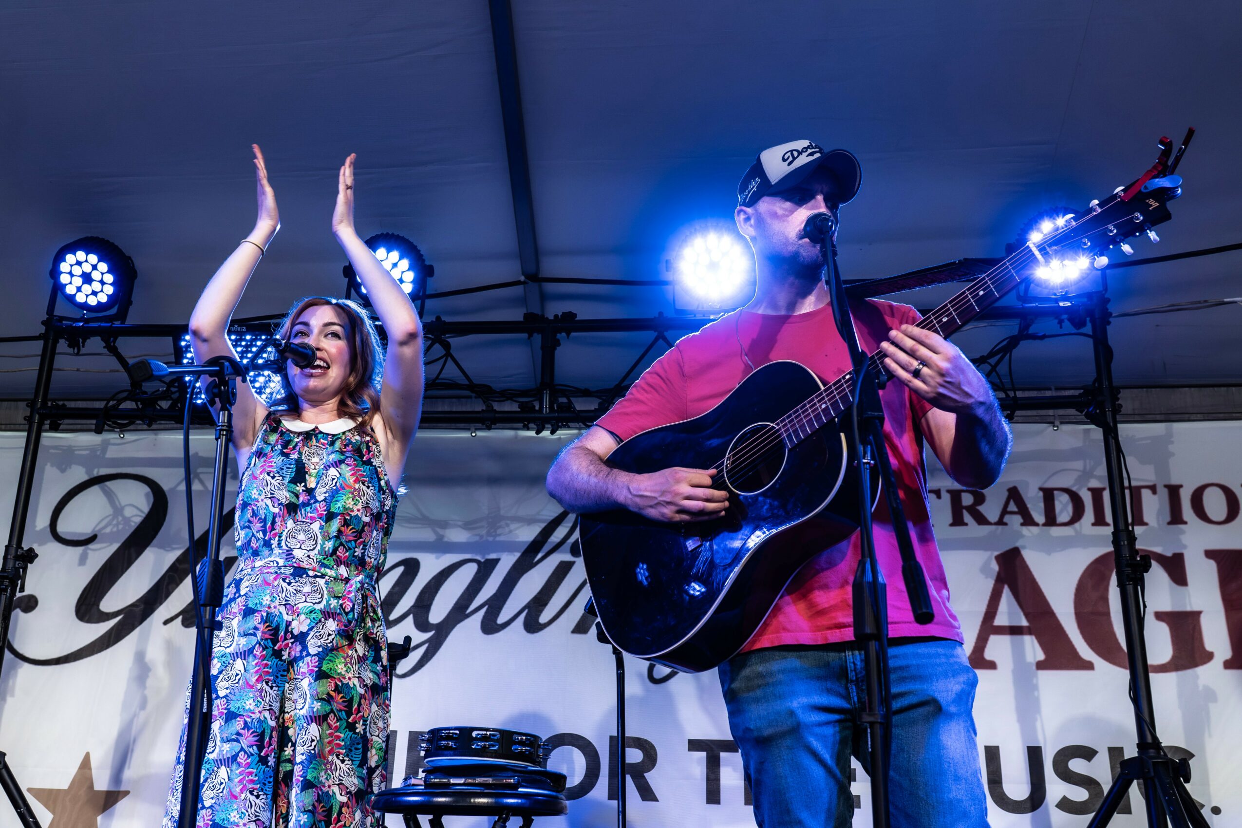 A woman and a man perform on stage under a tent, woman claps and man plays guitar with a microphone, stage lights visible.