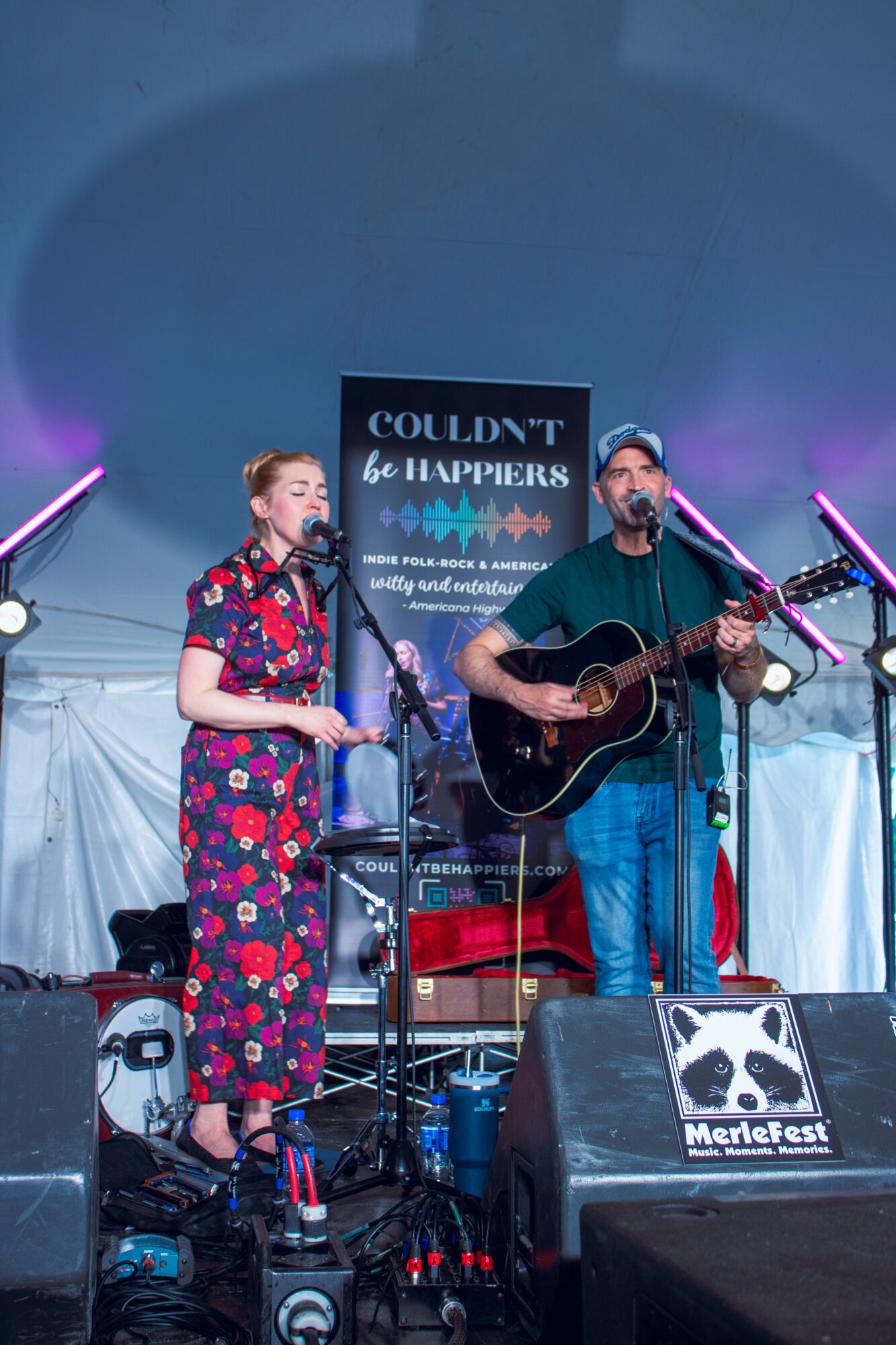 Two musicians perform on stage with a sign behind them reading 'COULDN'T be HAPPENS'. One plays guitar, the other sings, under a tent.