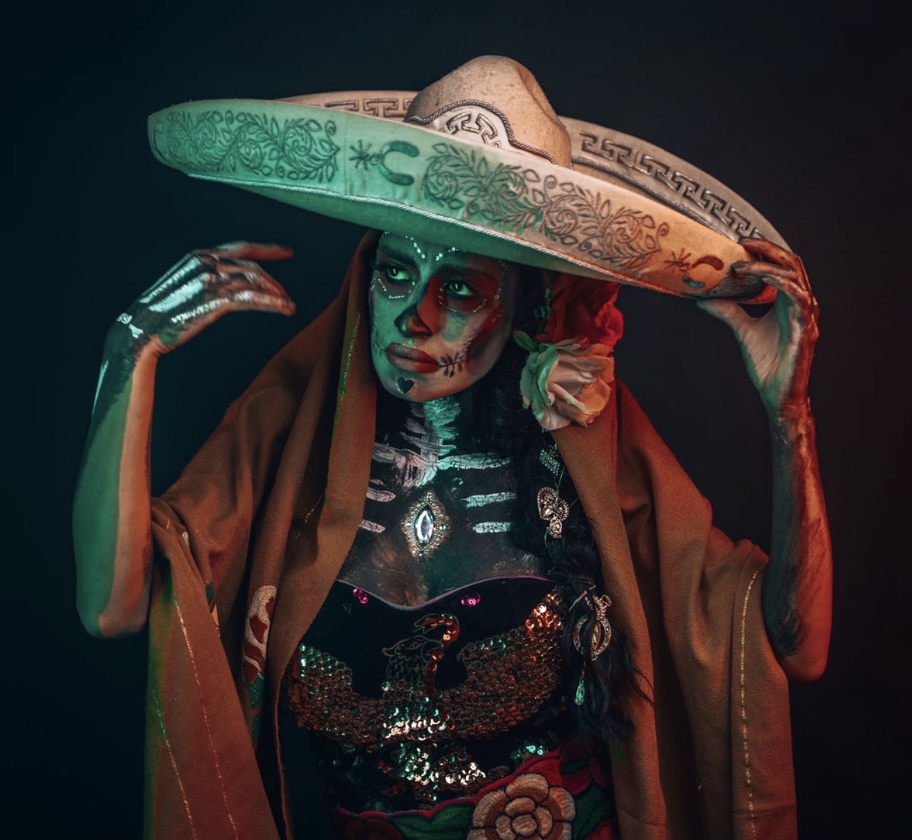 Person wearing a large sombrero, traditional clothing, and makeup with face paint, posing with hands near head, dark background.