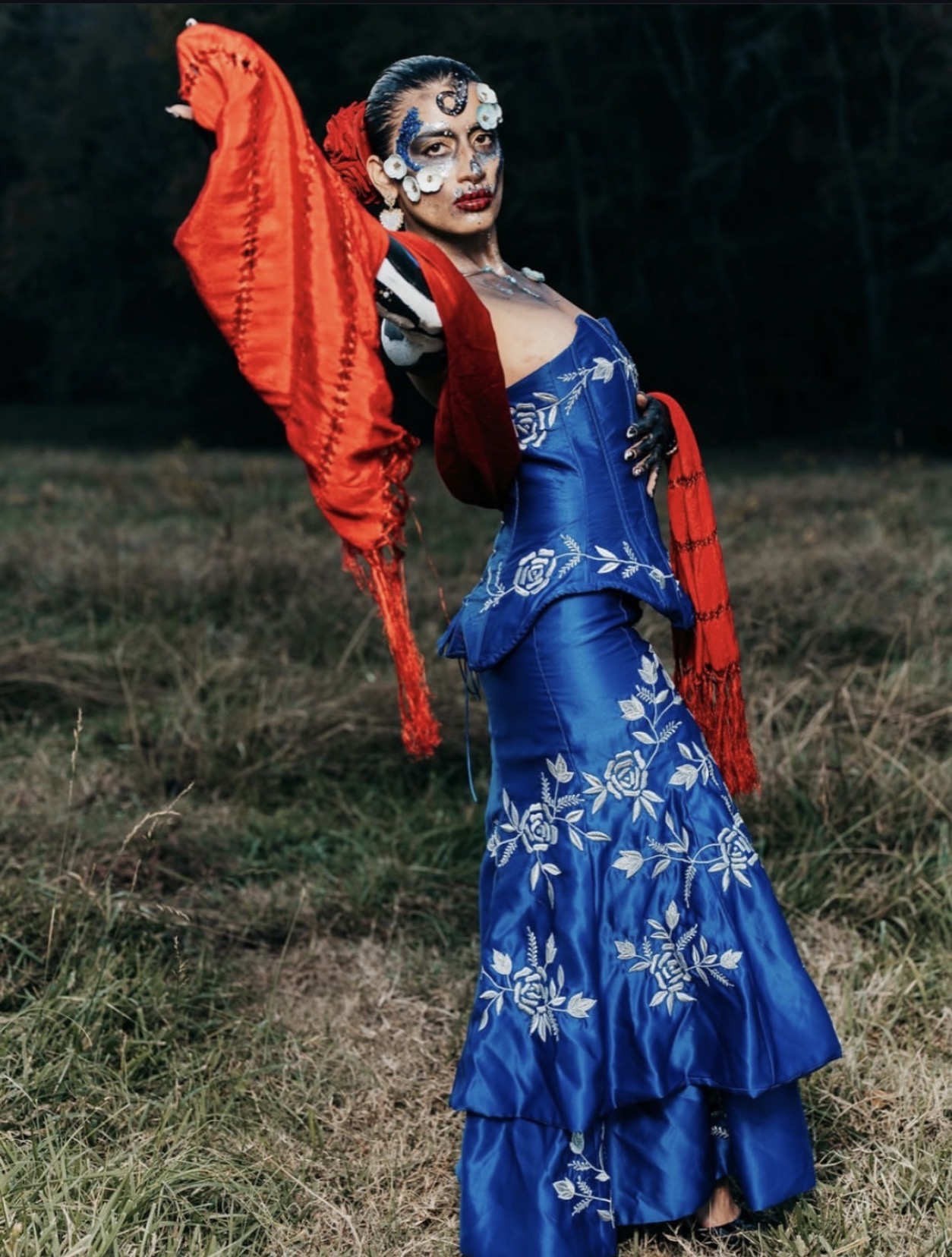 Person in colorful costume with face makeup, standing outdoors in a grassy area, holding a red fabric. Dark background.