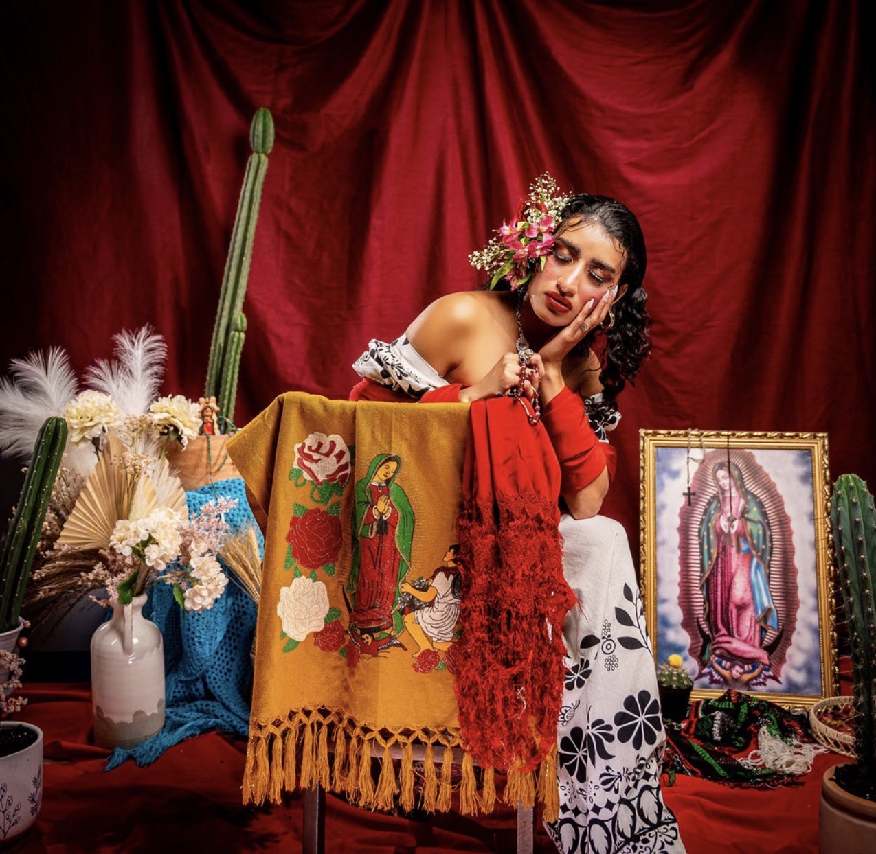 Woman with floral headpiece and traditional dress, surrounded by religious and decorative items, in front of red curtain.
