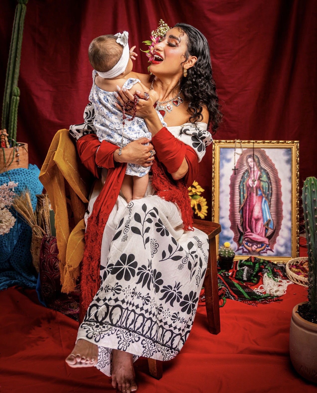 Woman holding a child, both wearing floral headbands, in front of a red curtain with religious artwork and plants nearby.