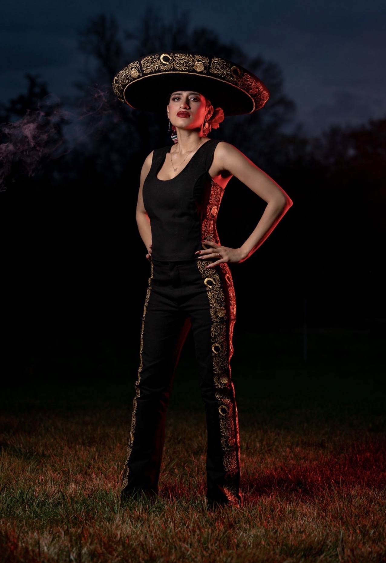 Woman in traditional Mexican attire with a large sombrero, standing outdoors at night with hands on hips.