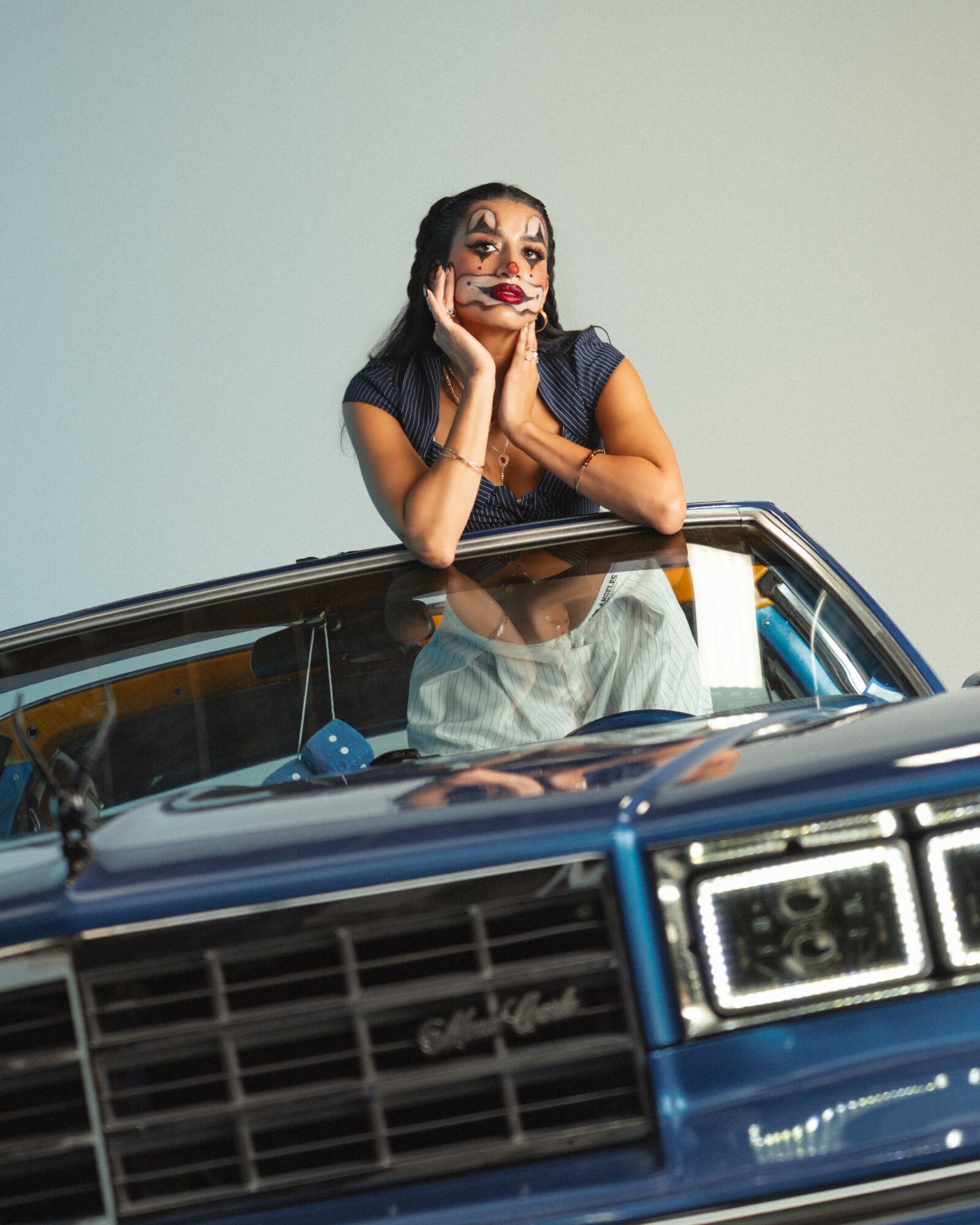 Person with clown makeup leaning on a blue car, resting chin on hands, smiling, against a plain background.