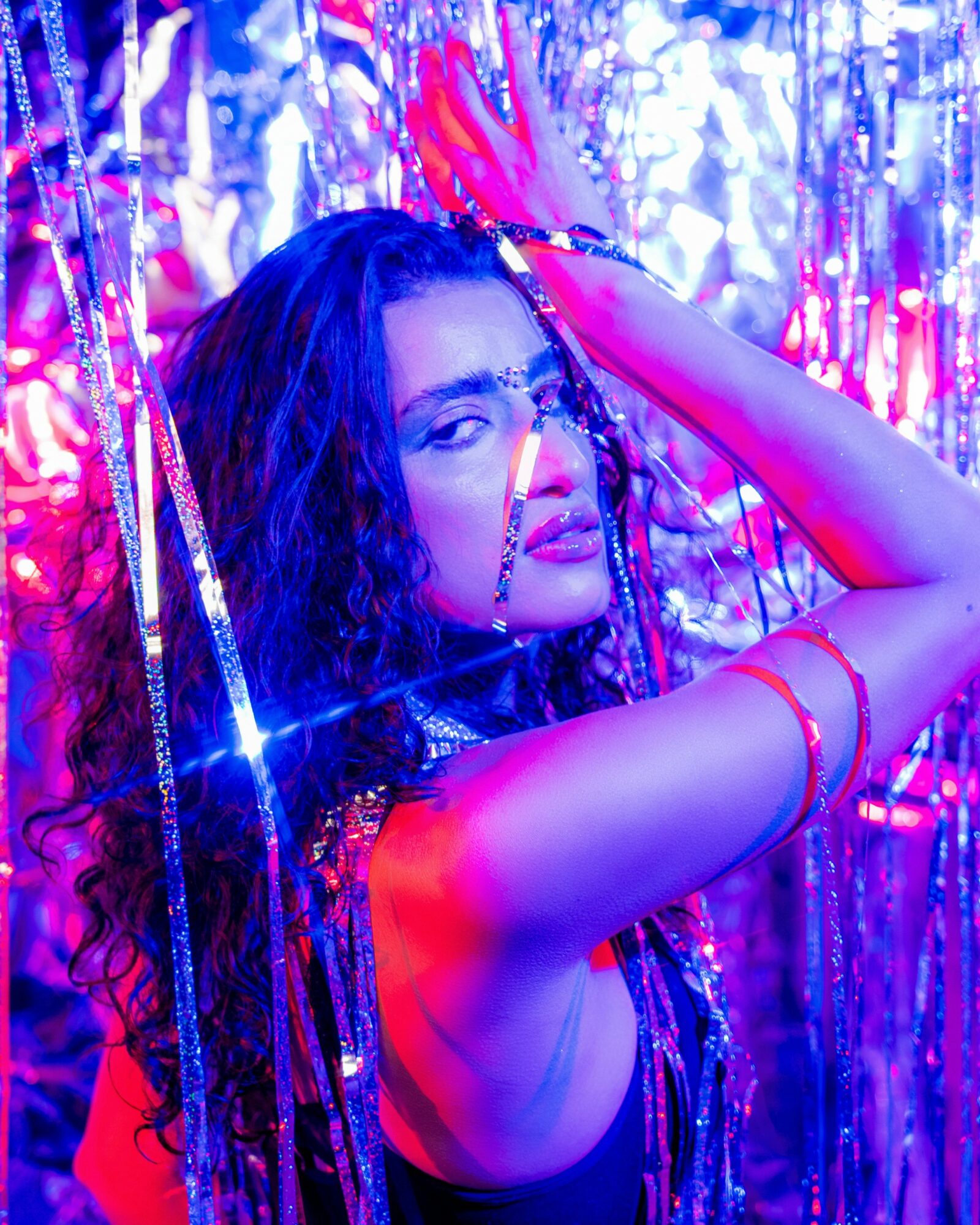 Woman with long curly hair poses with one arm raised, surrounded by shiny metallic streamers and colorful lighting.
