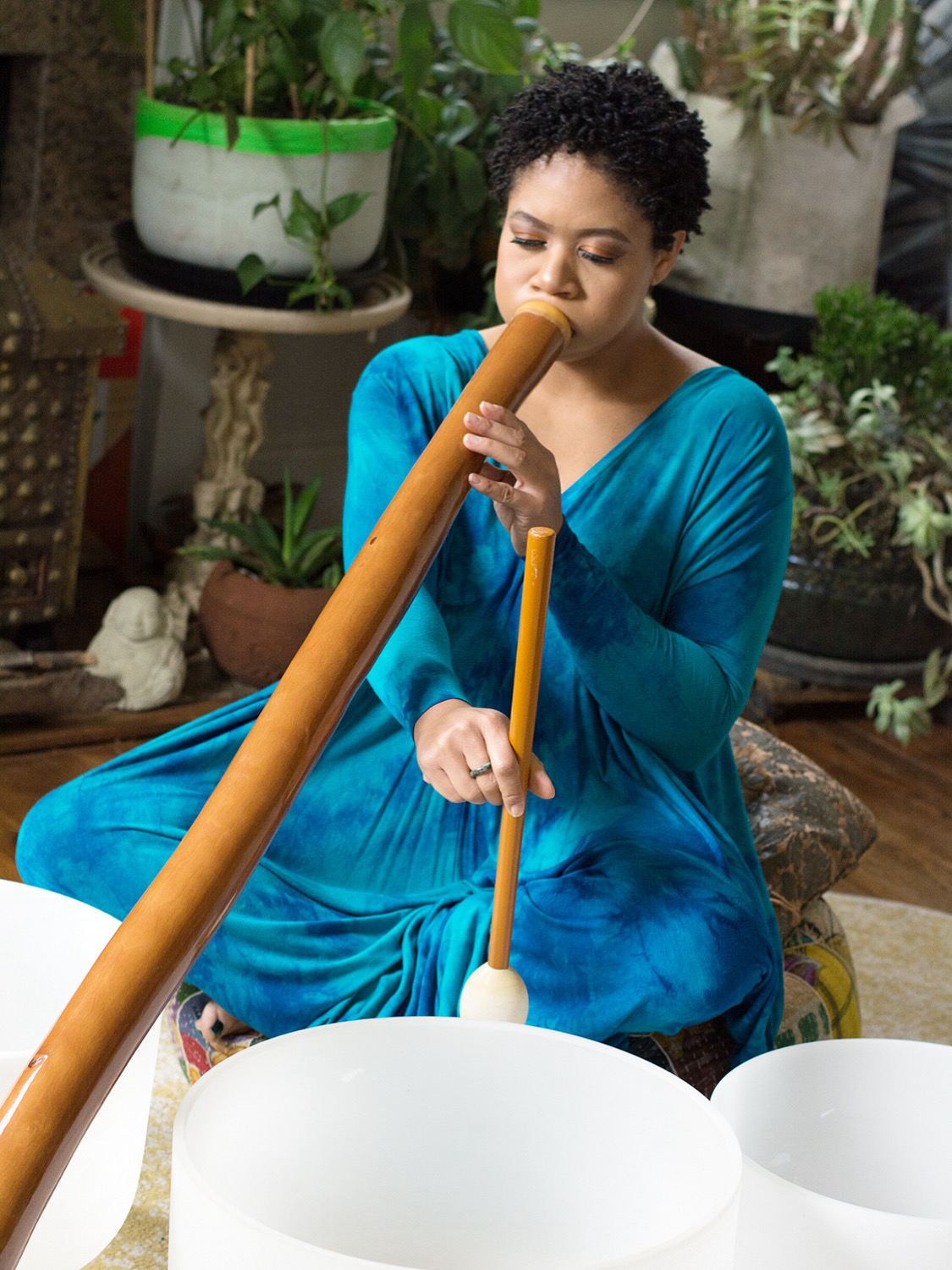 Woman in blue dress playing a didgeridoo indoors surrounded by plants and pottery.