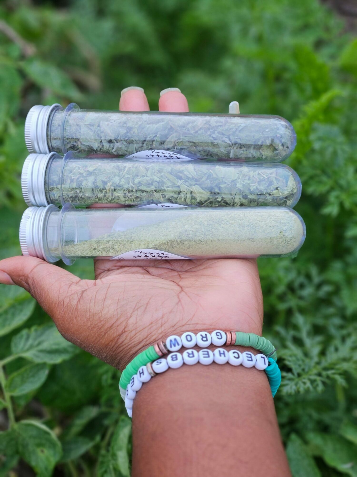 Person's hand holding three test tubes outdoors with green foliage in background, wearing a beaded bracelet.