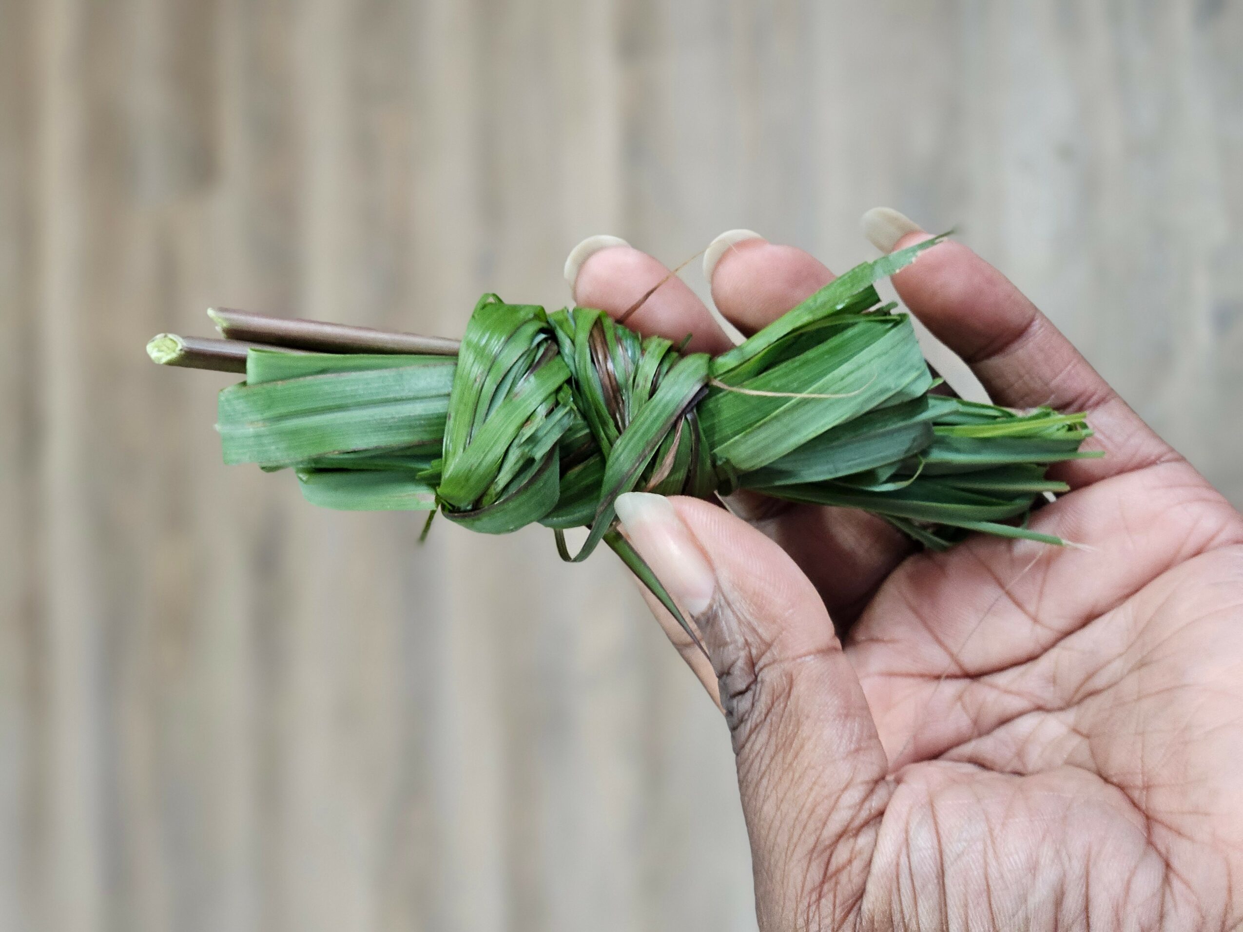 Hand holding a small bundle of green leaves tied together, with a wooden background.