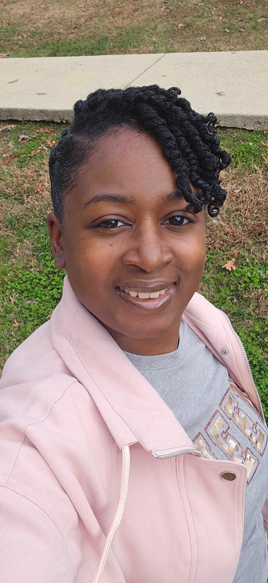 Young woman with curly hair smiling outdoors, wearing a light pink jacket and gray shirt.