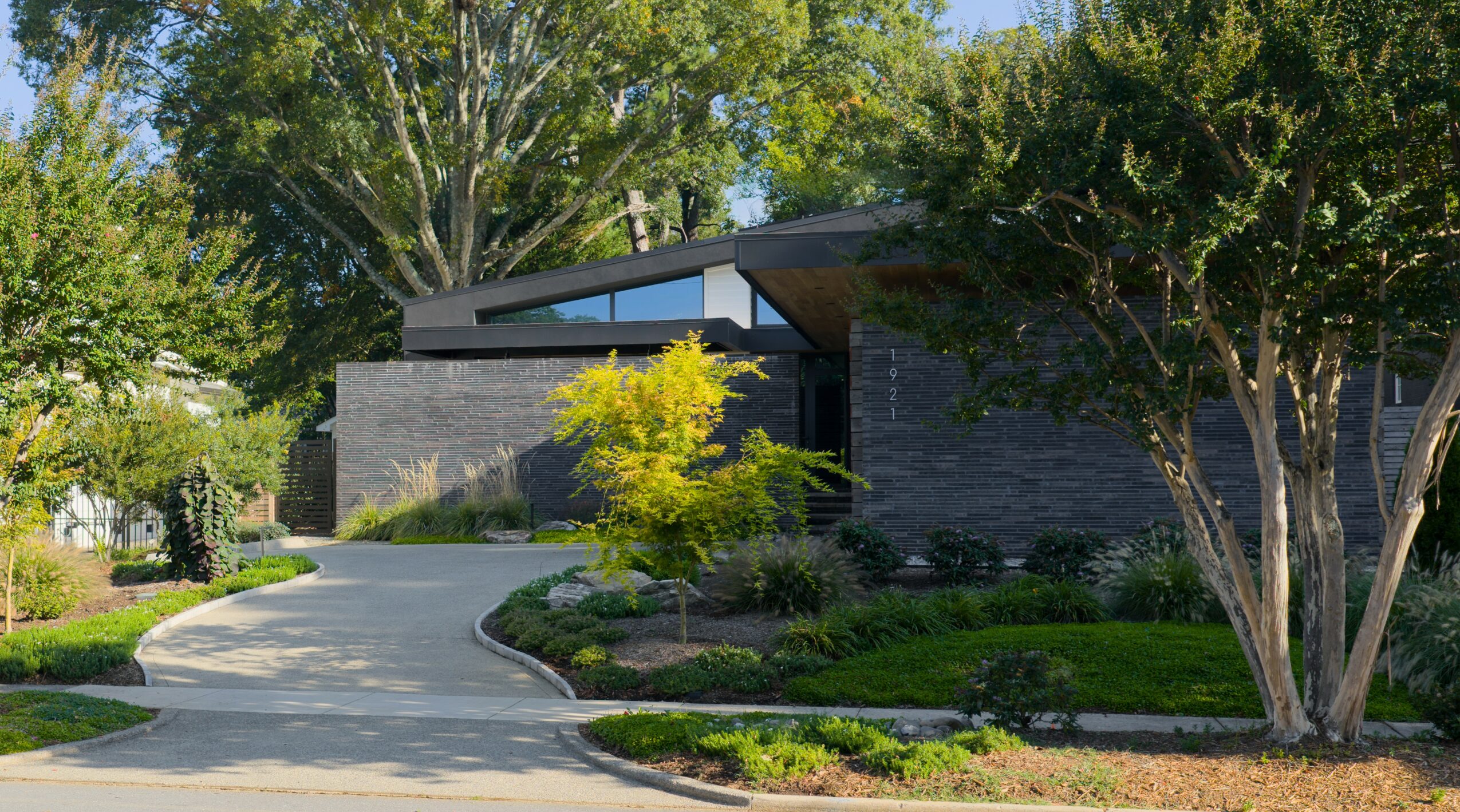 Modern house with a curved driveway, surrounded by trees and landscaped greenery, under a clear blue sky.