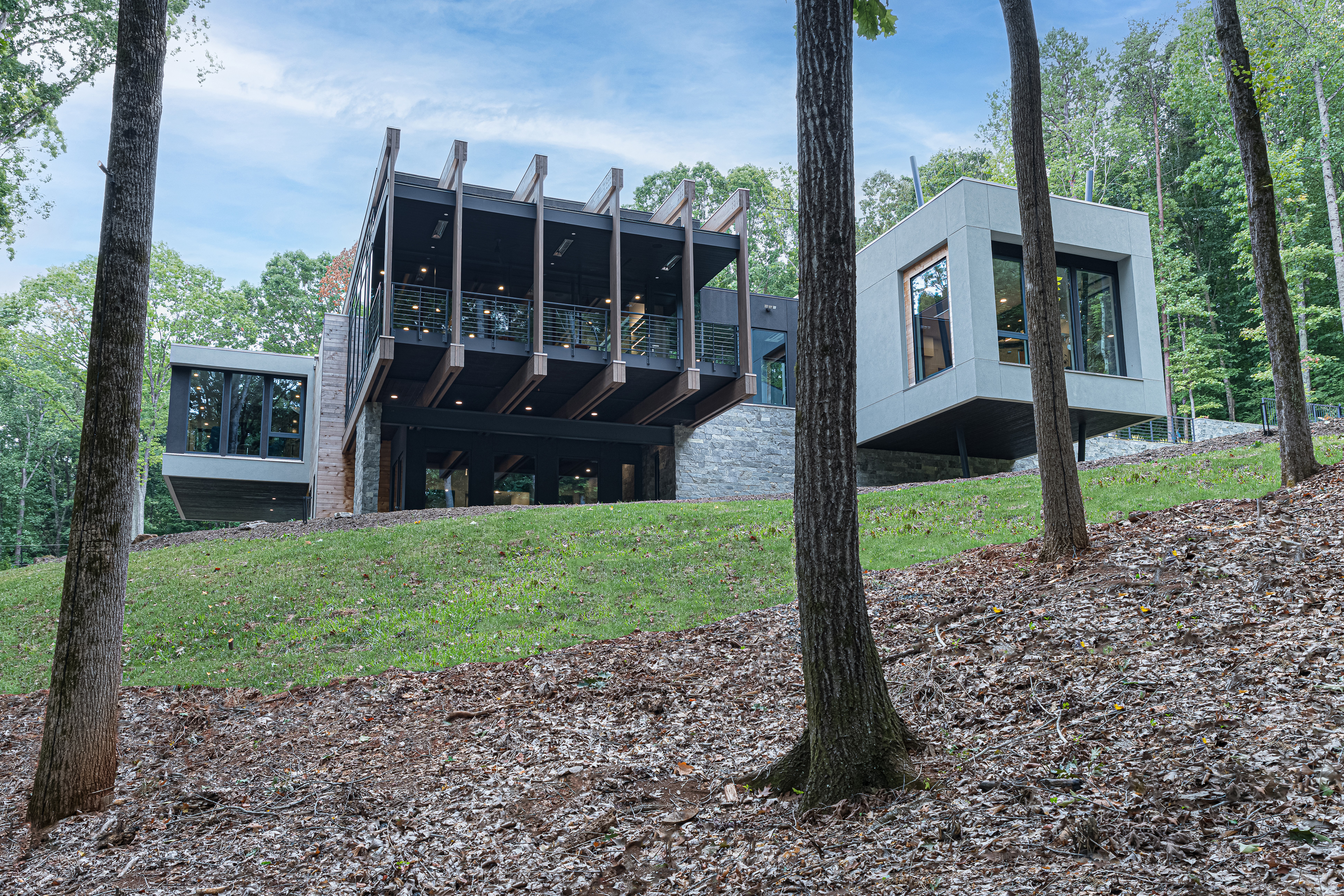Modern house with large windows and a balcony, situated on a grassy hillside surrounded by trees.