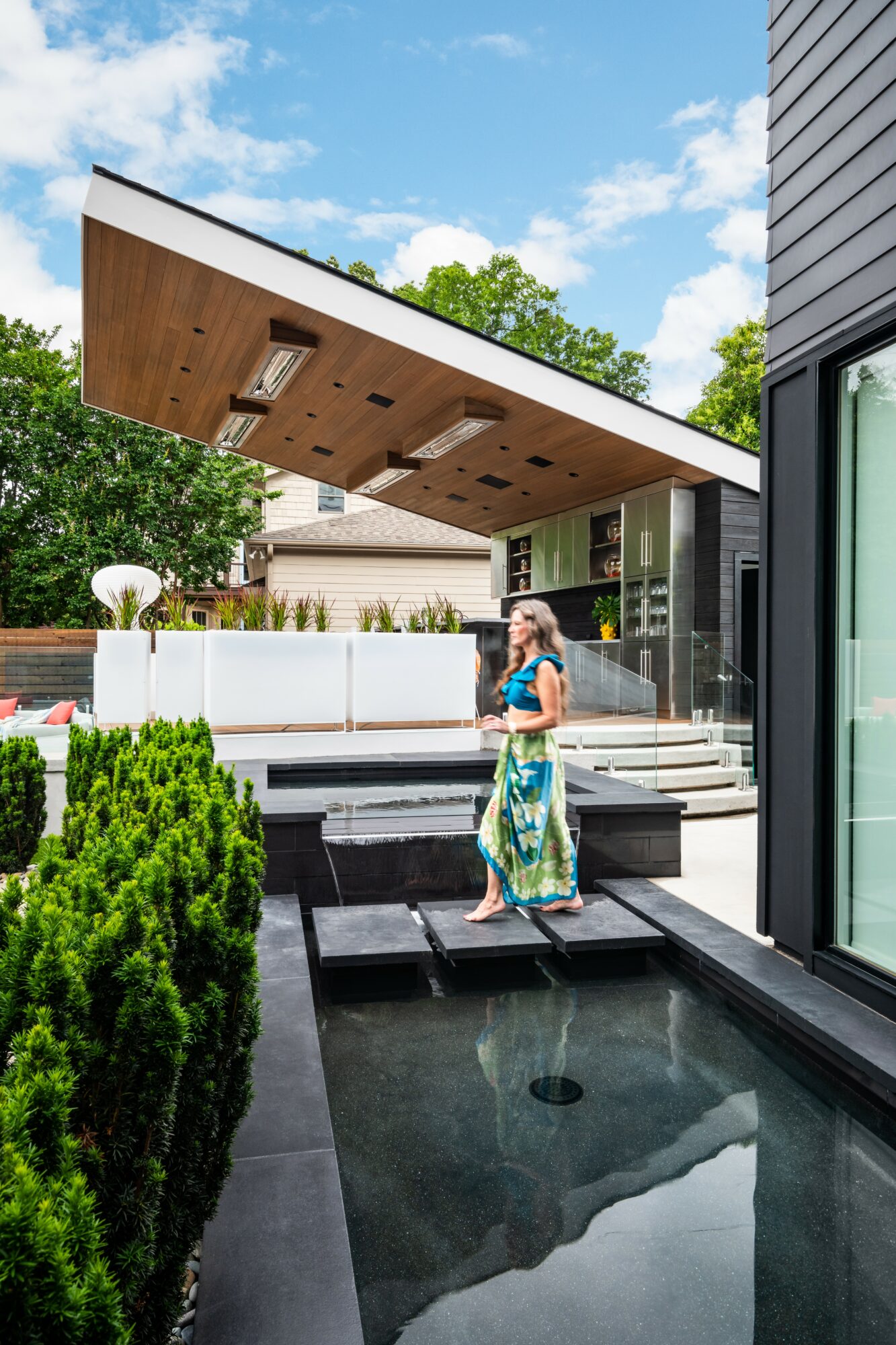 Woman walking on a modern outdoor patio with water feature, greenery, and contemporary building architecture under a blue sky.