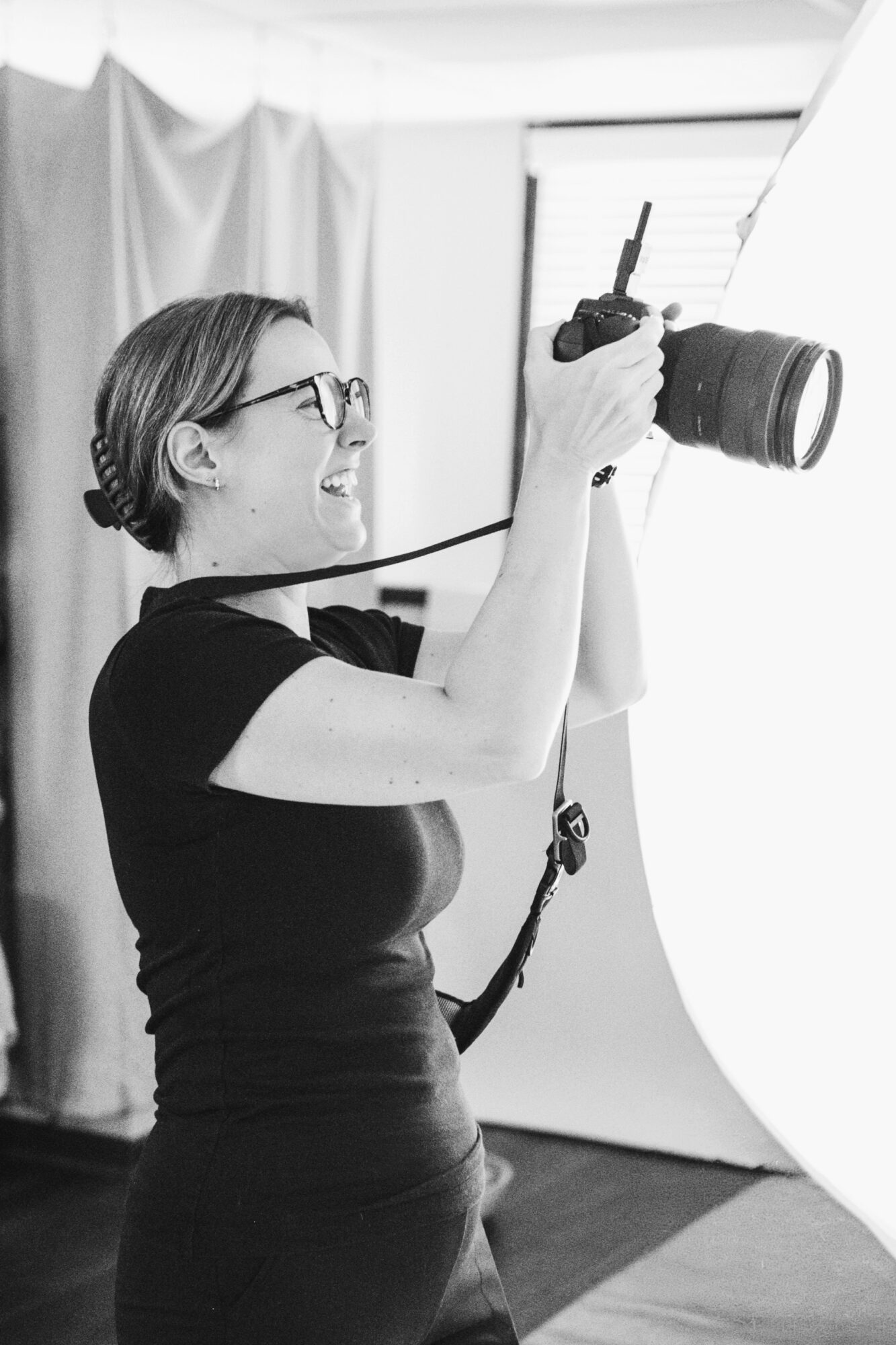 Woman with glasses holding a camera, smiling, in a photography studio with a white backdrop