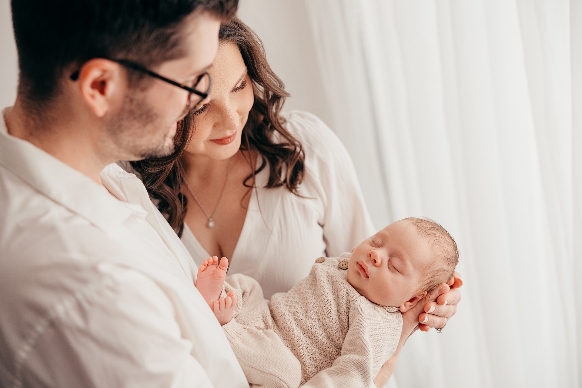 A couple holding a sleeping baby near a window with white curtains, soft natural light, and gentle expressions.