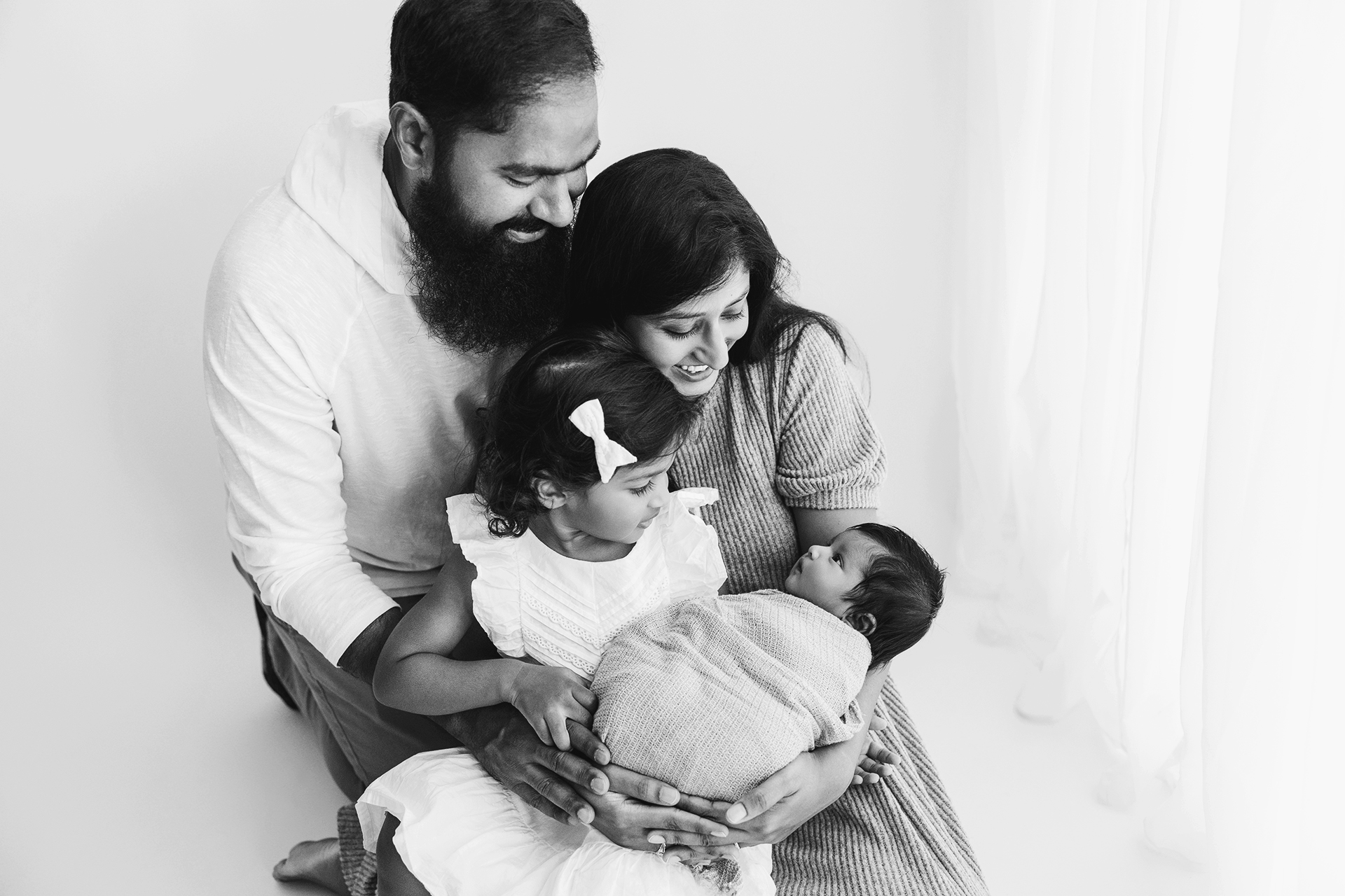 Family of four with a man, woman, young girl, and baby, smiling and looking at the baby, in a bright room.