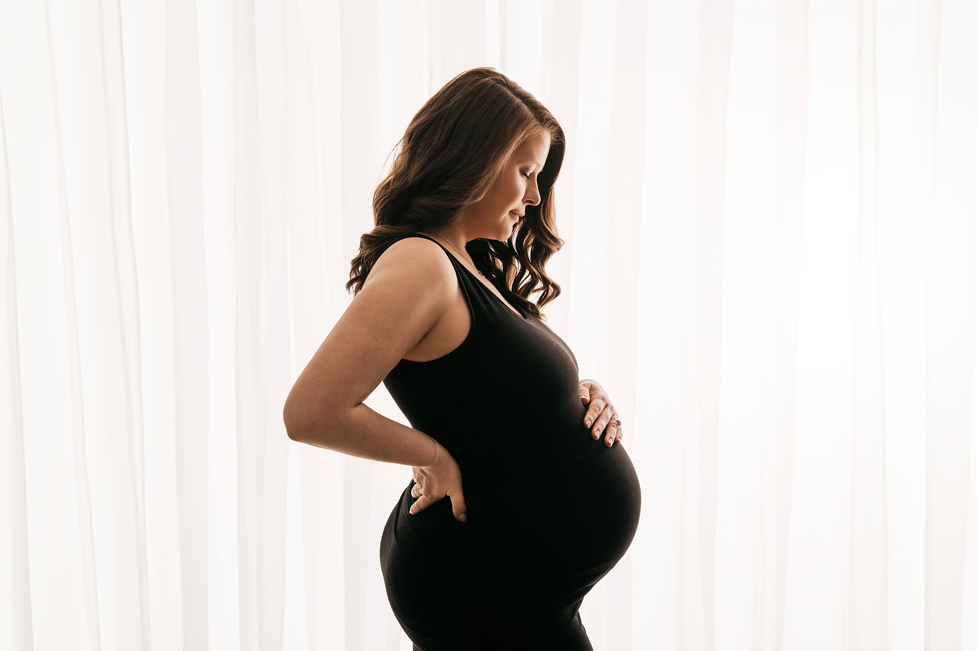 Pregnant woman standing sideways with hands on her belly, in front of white curtains, wearing a black sleeveless top.