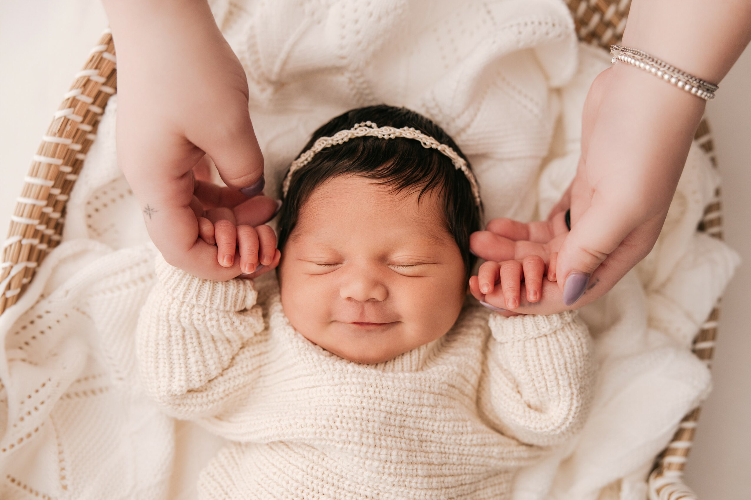 Baby with eyes closed, wearing a cream sweater and headband, lying on a white blanket, holding hands with two adults.