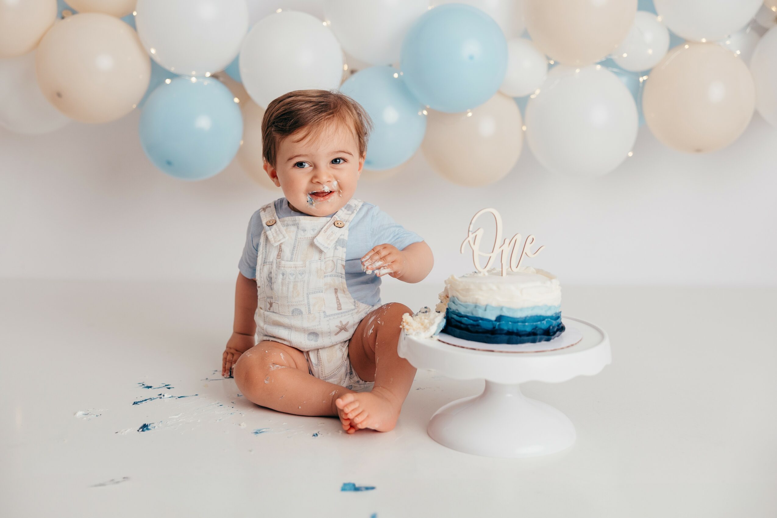 Smiling toddler sitting on floor with balloons in background, next to a cake on a stand, celebrating a first birthday.