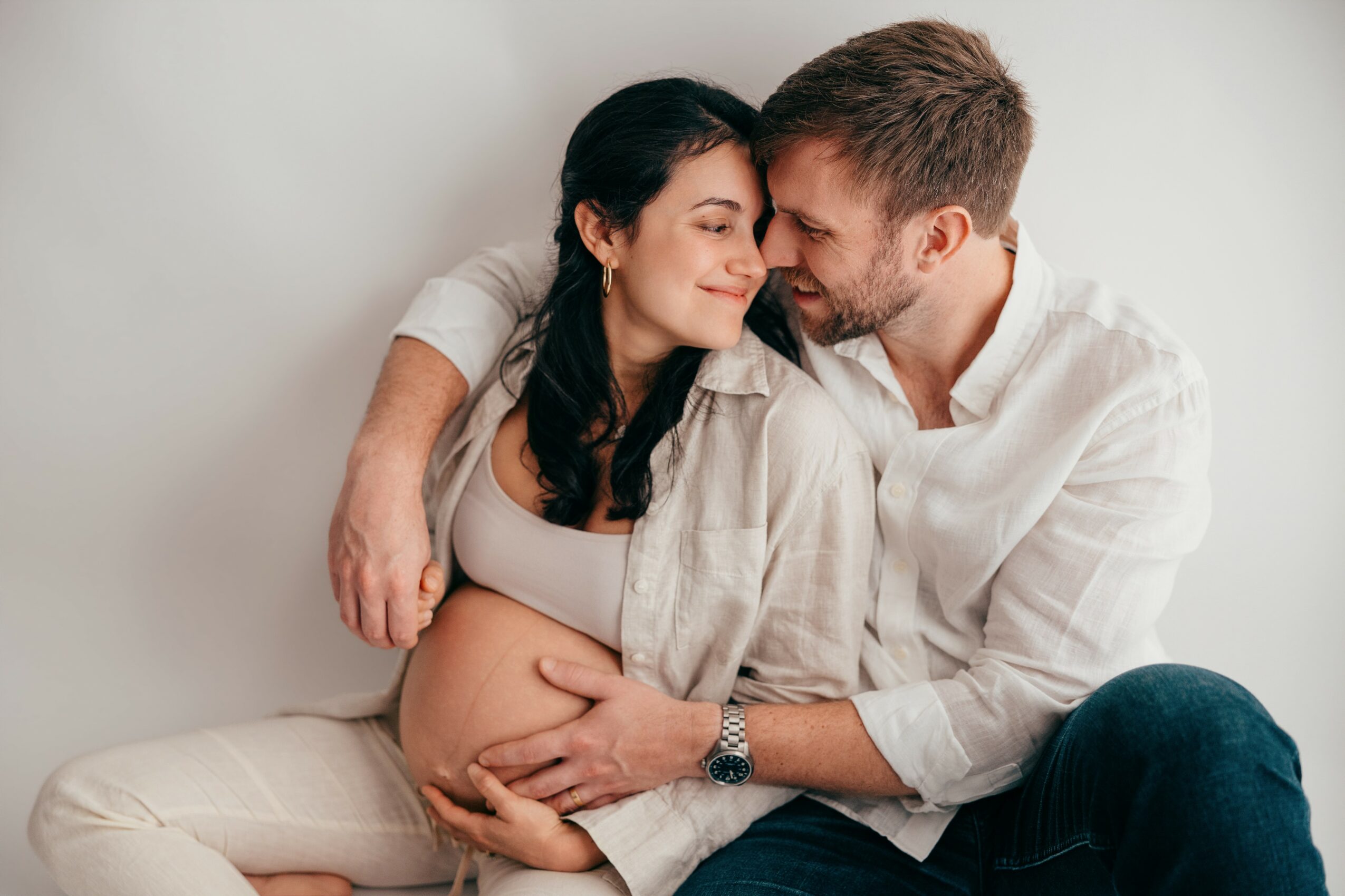 Pregnant woman and man sit close, touching her belly, smiling, in a cozy setting.