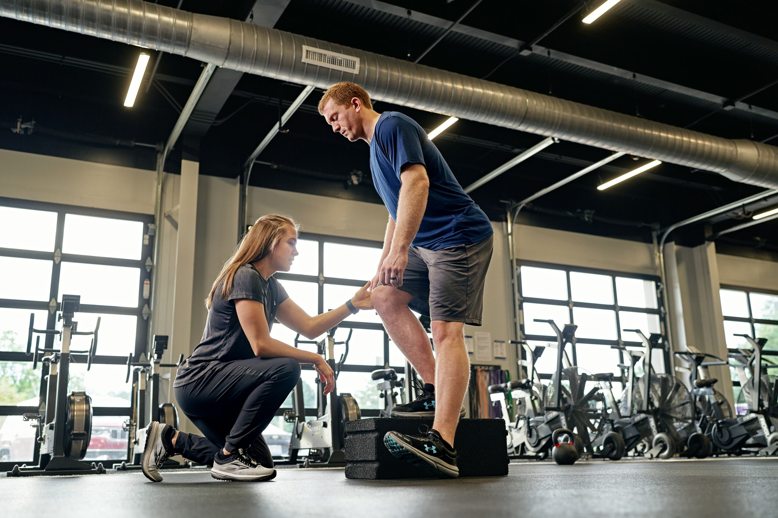 A trainer assists a woman with her workout in a gym, holding her hand as she stands on a step platform.