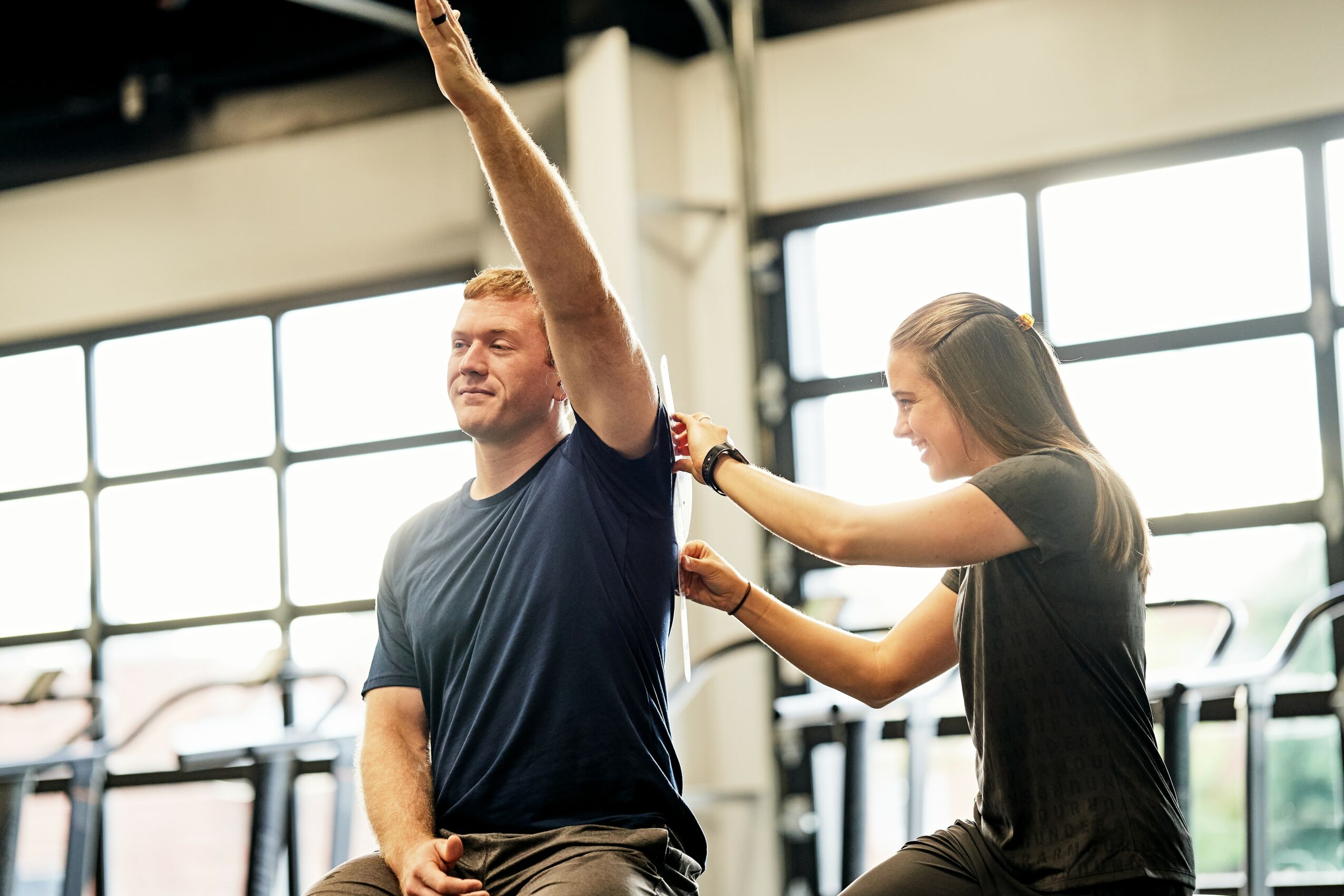 A man and woman stretch their arms in a gym with large windows and exercise equipment in the background.