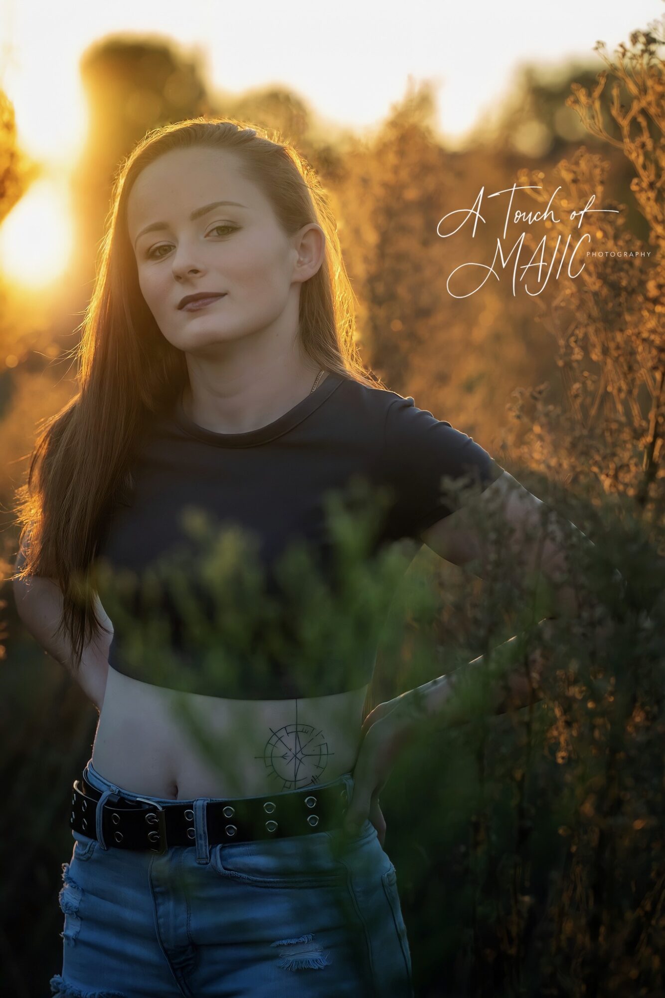 Young woman with long hair outdoors during sunset, wearing a dark top and jeans, with greenery in foreground.