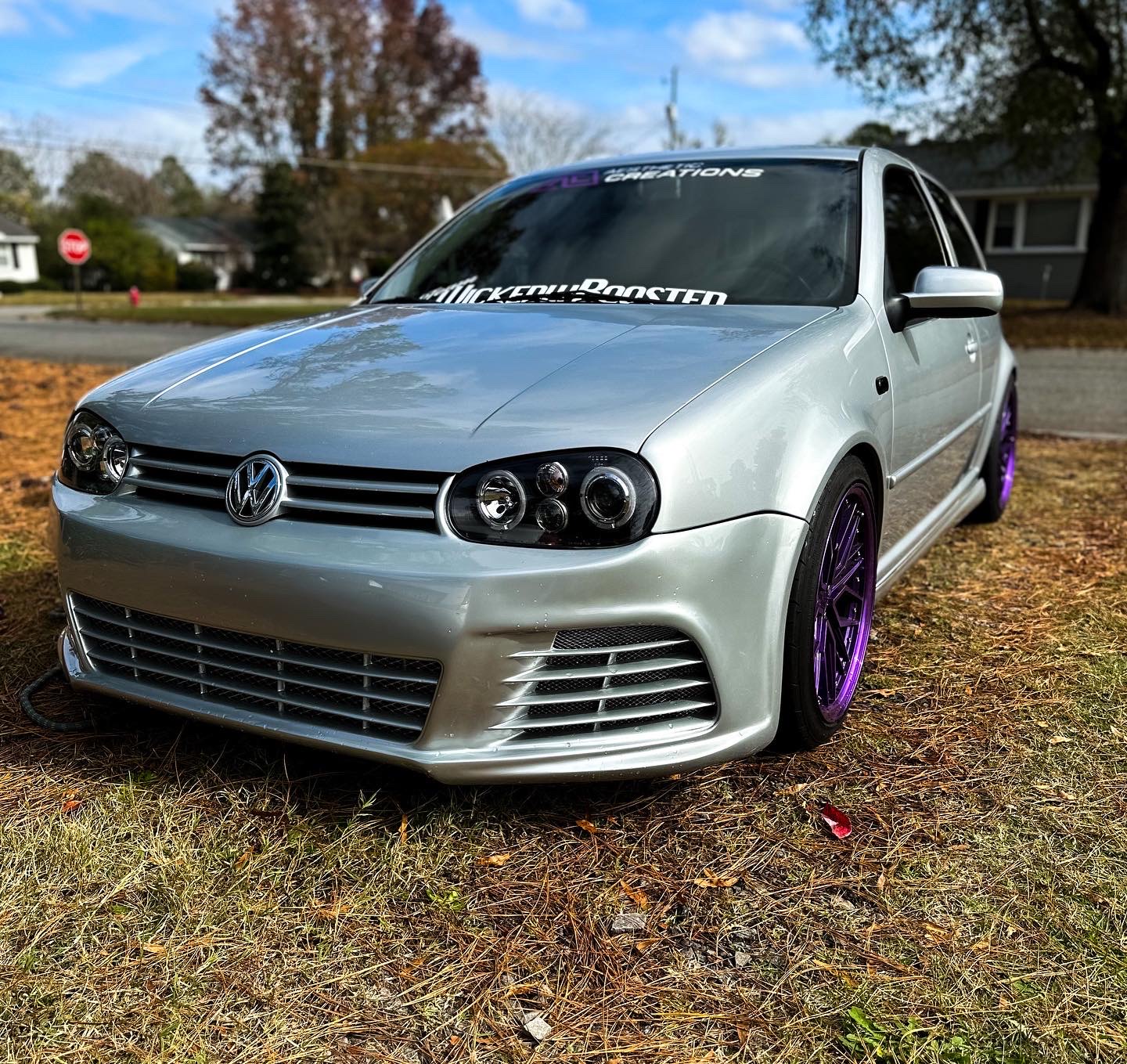 Silver Volkswagen car with purple wheels parked on grass, trees and houses in background, sunny day.