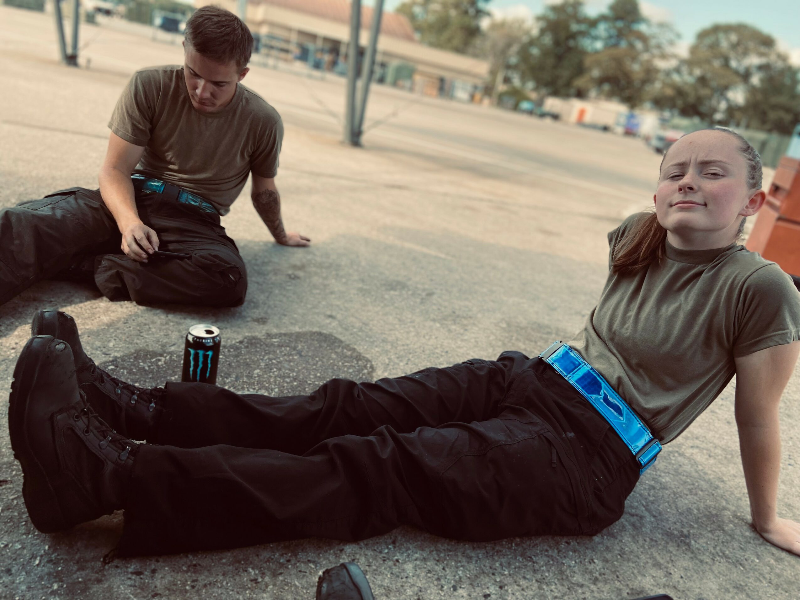 Two people sitting on the ground outdoors, one leaning back with legs extended, the other sitting nearby, with a clear sky and trees in background.