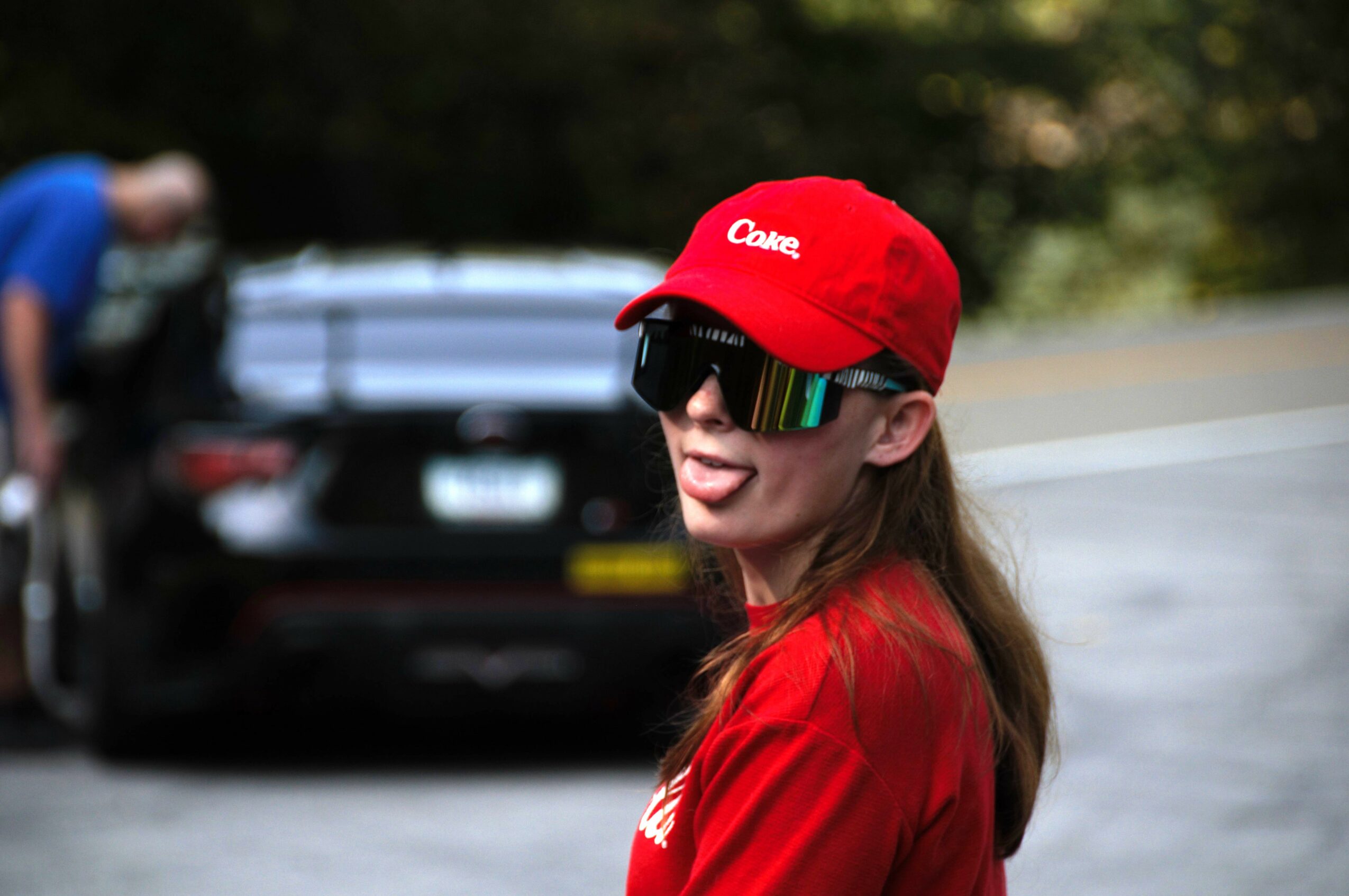 Woman wearing red cap and sunglasses smiling outdoors, with a black car and trees in the background.