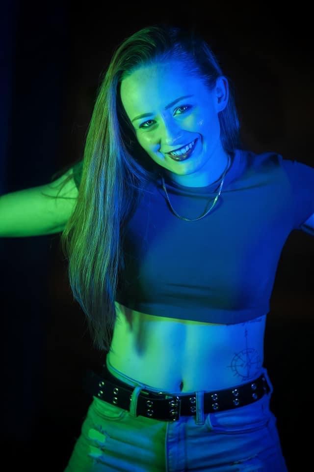 Young woman smiling, with long hair, wearing a crop top and jeans, illuminated by colorful lighting.