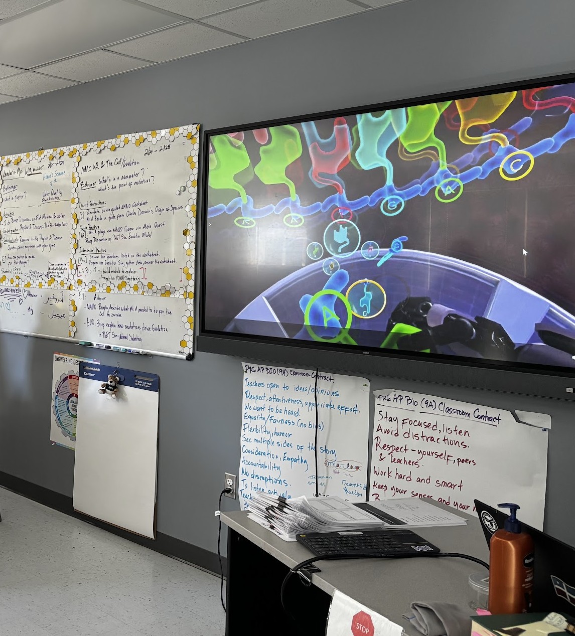 Indoor room with whiteboards, a monitor displaying colorful graphics, and a desk with papers and a water bottle.