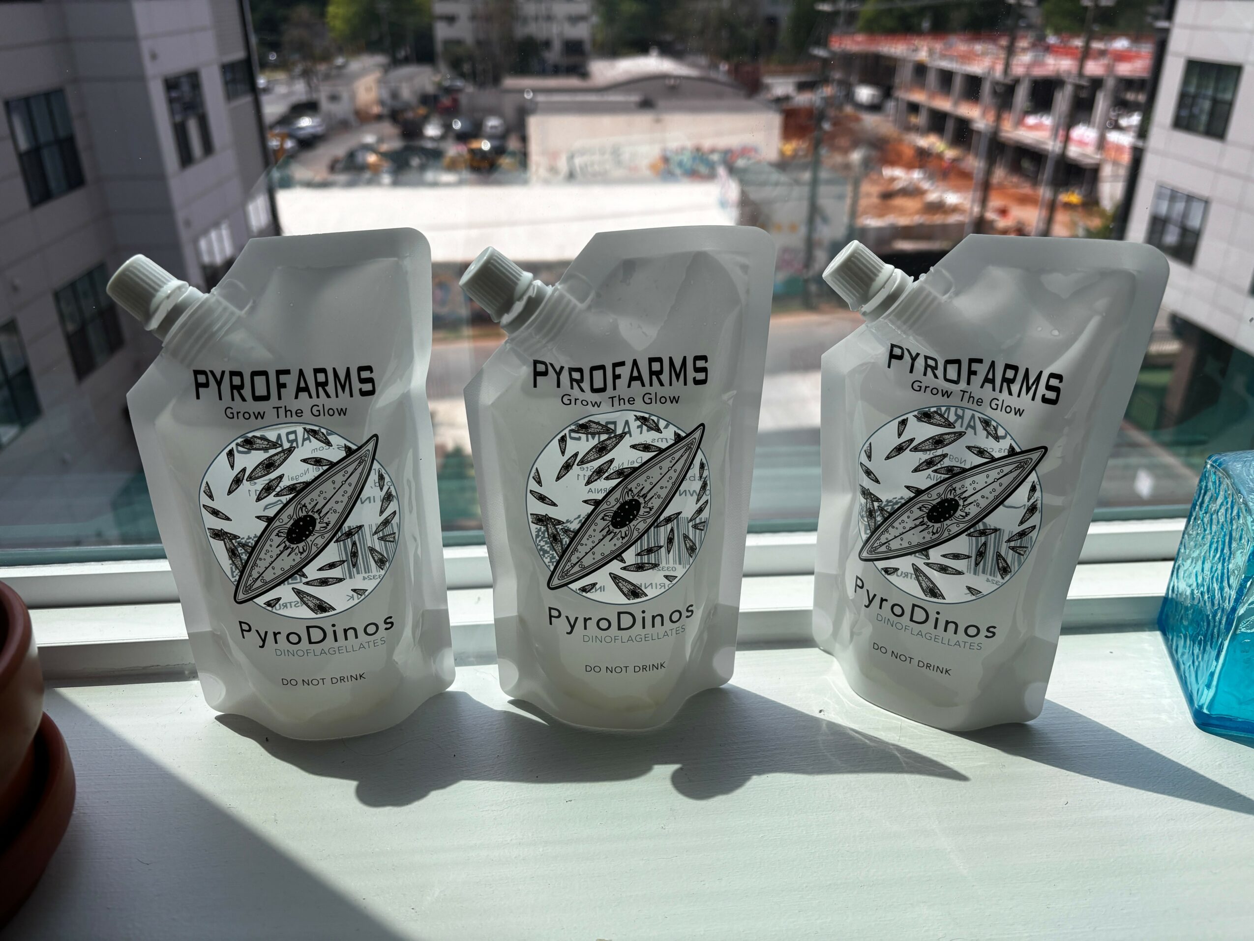 Three white pouches with a logo of a dinosaur and a leaf, placed on a windowsill with city view in background.