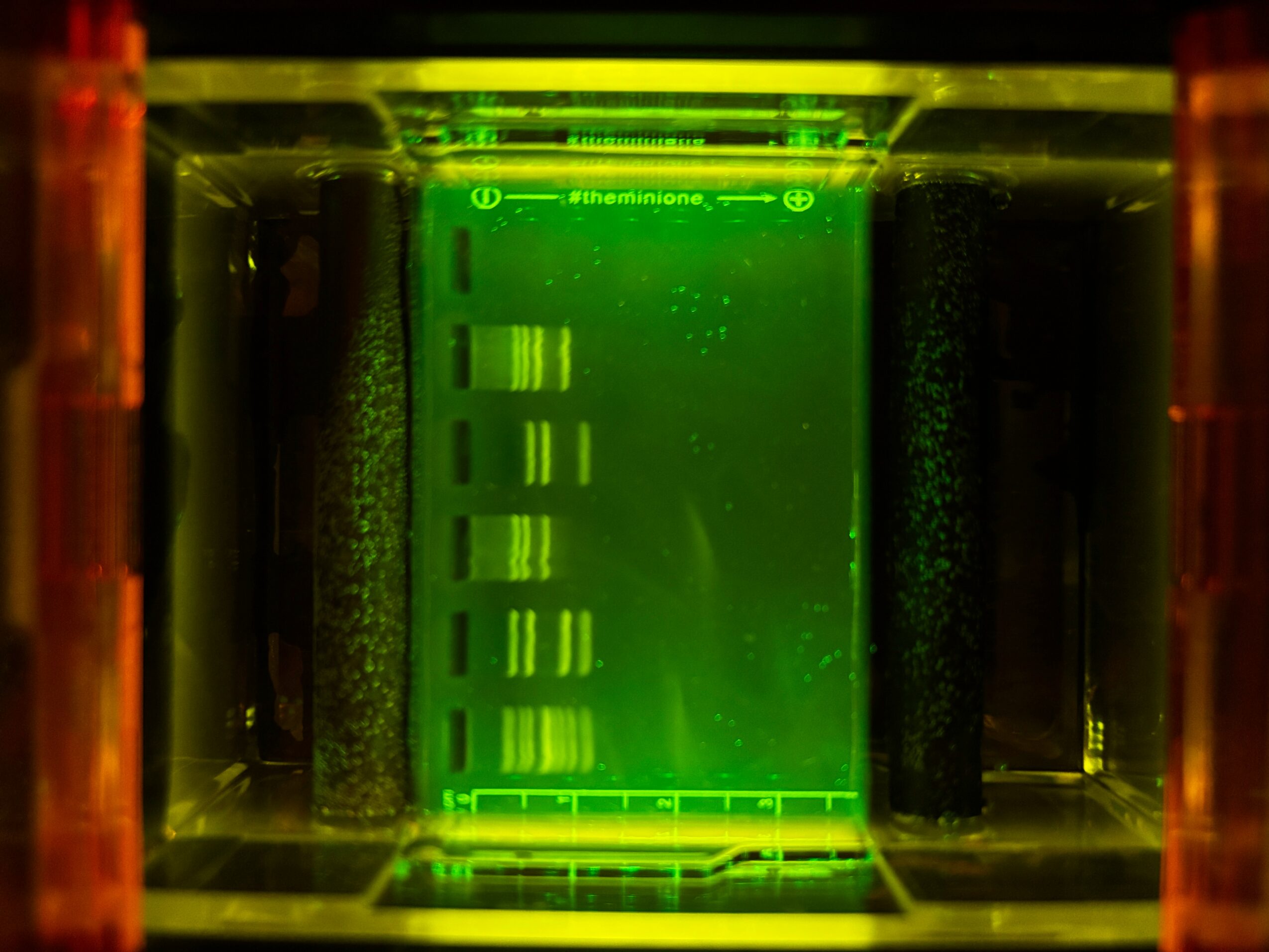 View inside a scientific device with green illumination, vertical black cylinders, and a central green-lit panel with markings.