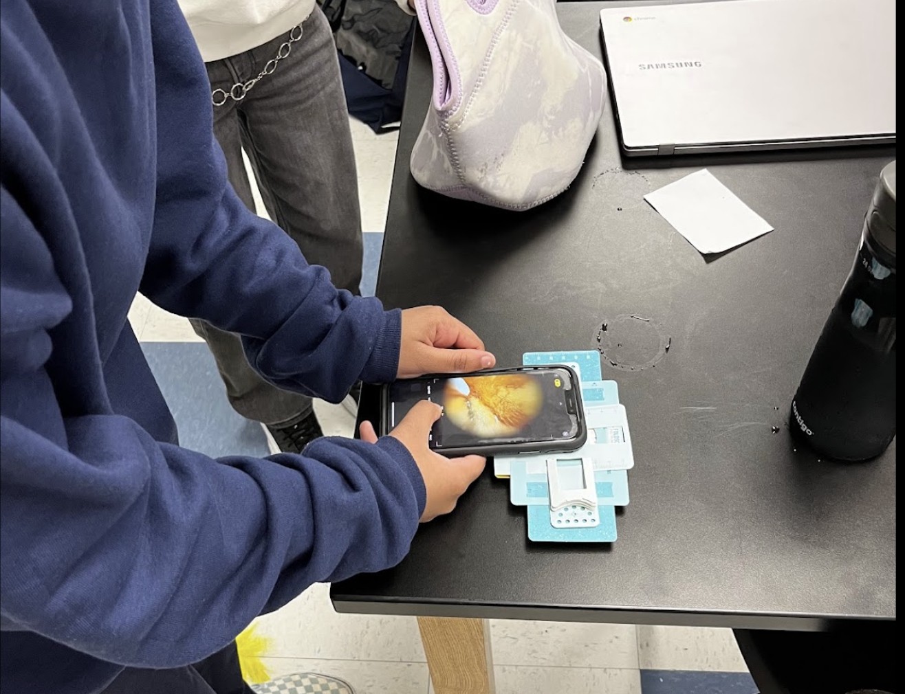 Child in blue sweatshirt holding a smartphone over a black table with various objects, including a white bag and a laptop.
