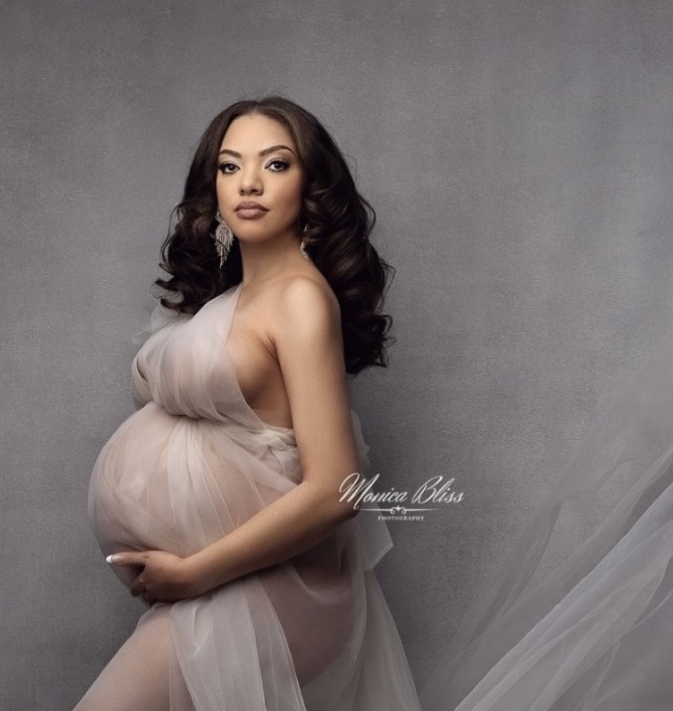 Pregnant woman with dark hair in loose curls, wearing a sheer, flowing dress, holding her belly, standing against a gray background.