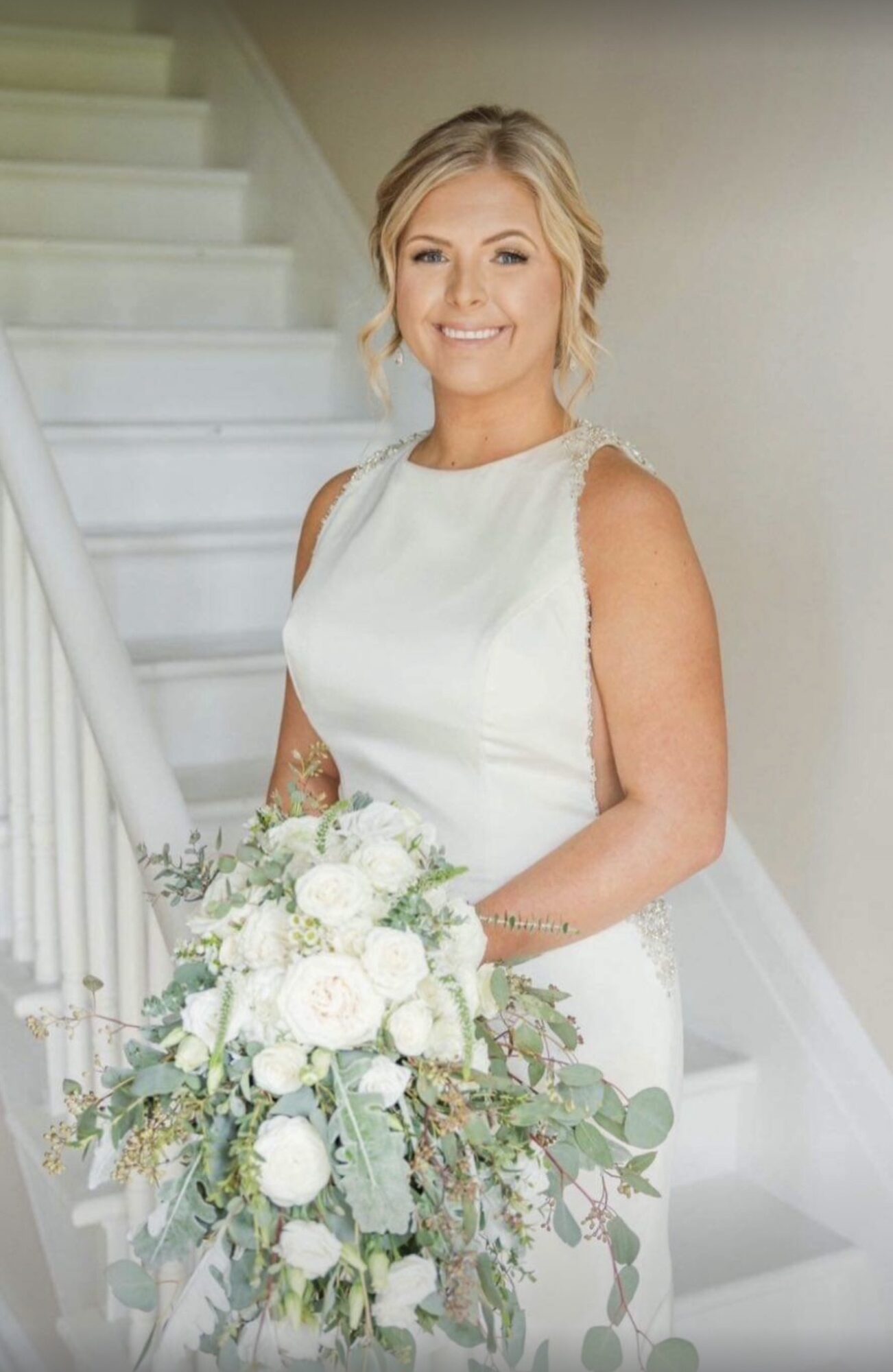 Smiling woman in wedding dress holding a large bouquet of white flowers, standing on a staircase.